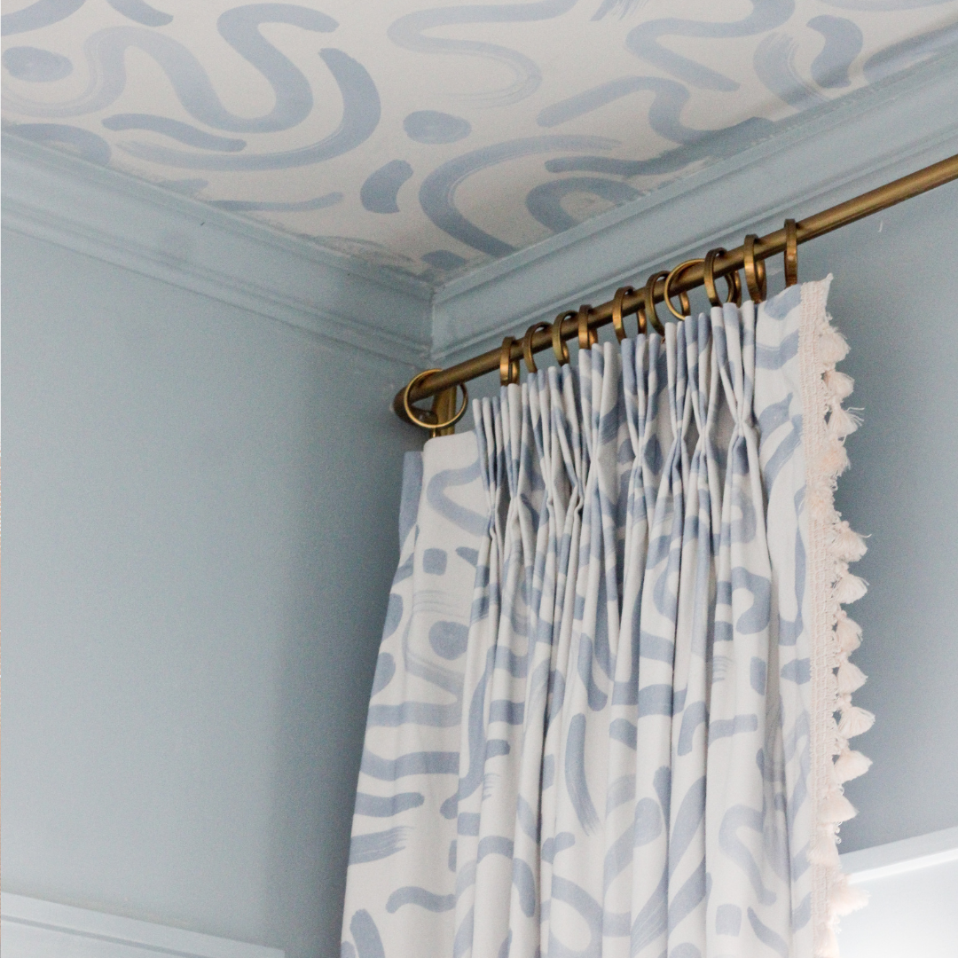 Close up of the top of a whimsical blue curtain with cream tassel trim hanging on a brass rod, styled with blue whimsical wallpaper on the ceiling