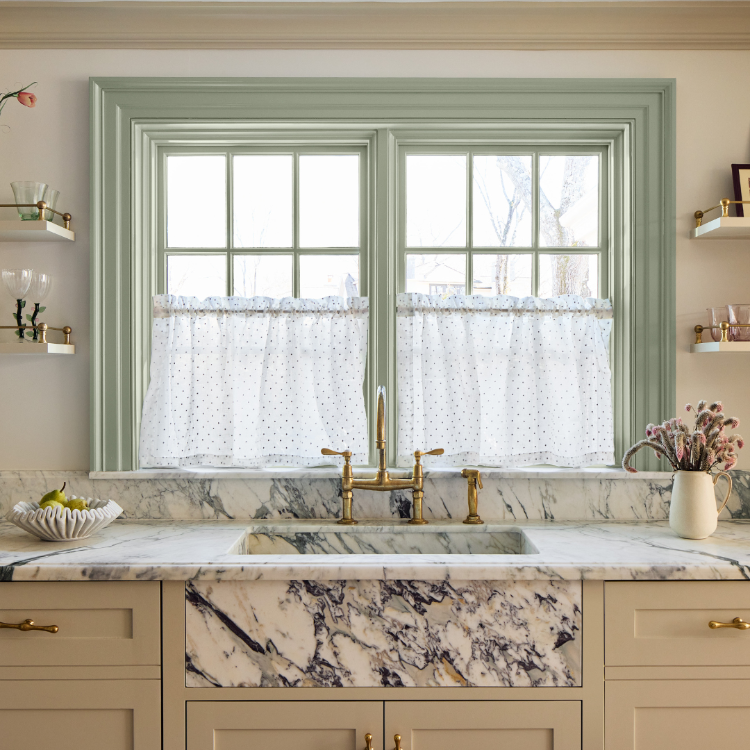 Close up of a black and white marble kitchen sink with a window behind it covered with sheer cafe curtains and green painted window trim