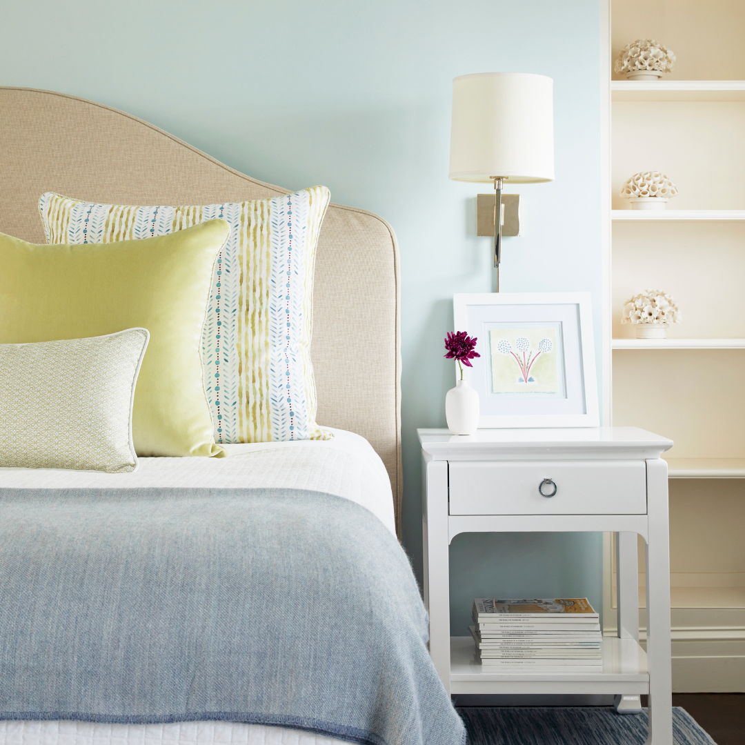 Close up of blue painted bedroom with a tan headboard styled with blue and green striped custom pillow, light green custom pillow, and blue and green geometric patterned pillow next to a white nightstand with floral artwork