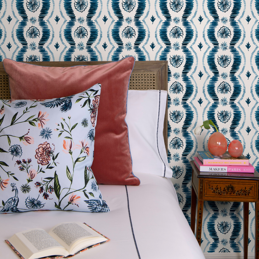 Bedroom vignette with blue ikat clay coated wallpaper and a wooden bed frame styled with white sheets and a coral velvet pillow and blue chinoiserie pillow