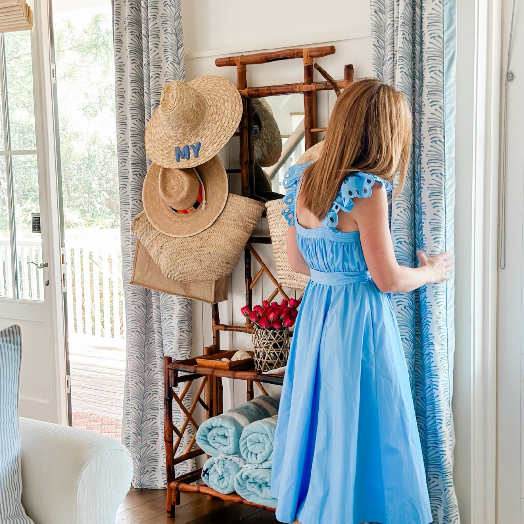 Woman wearing a long blue dress holding Sky Blue Botanical Stripe custom curtains next to a wooden hanger stand with beach hats, totes, and towels