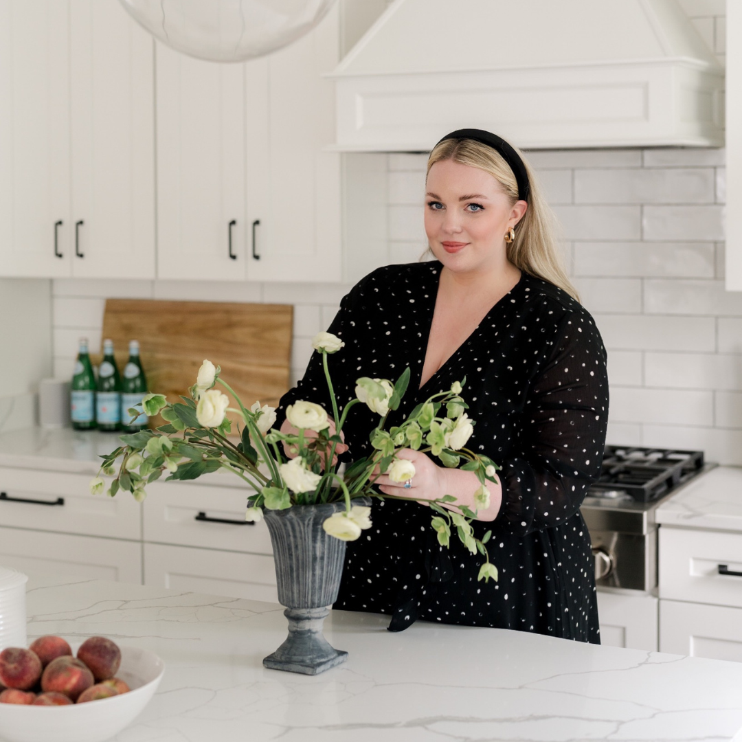 Blonde woman standing in a white kitchen holding flowers in grey vase on top of white granite counter