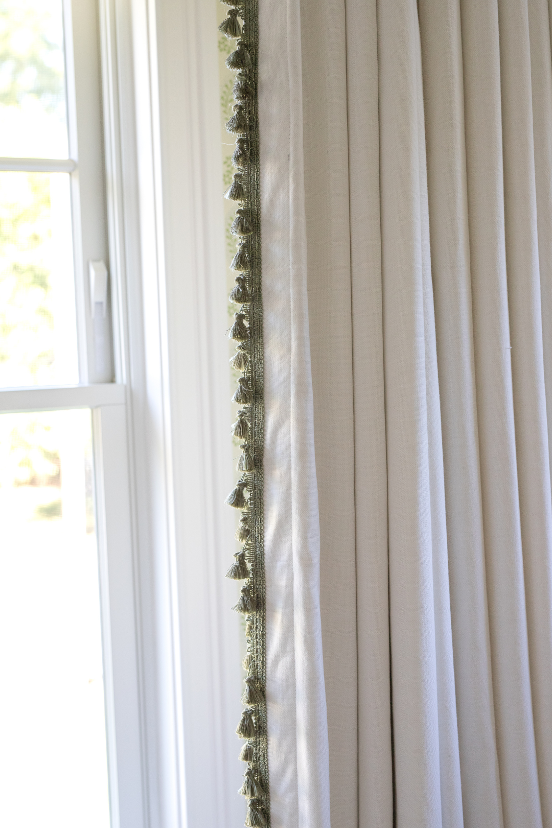 Natural while curtain with green tassel trim in front of a window panel