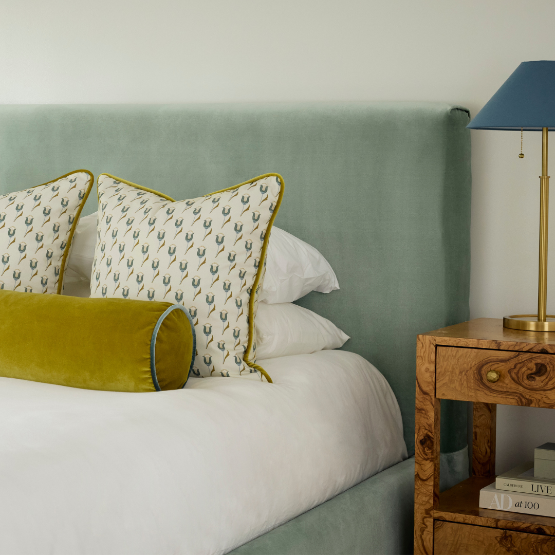 Close up of a king bed upholstered in blue green velvet paired with white sheets, blue and green tulip patterned pillows and a citron bolster pillow. next to a wooden sidetable.