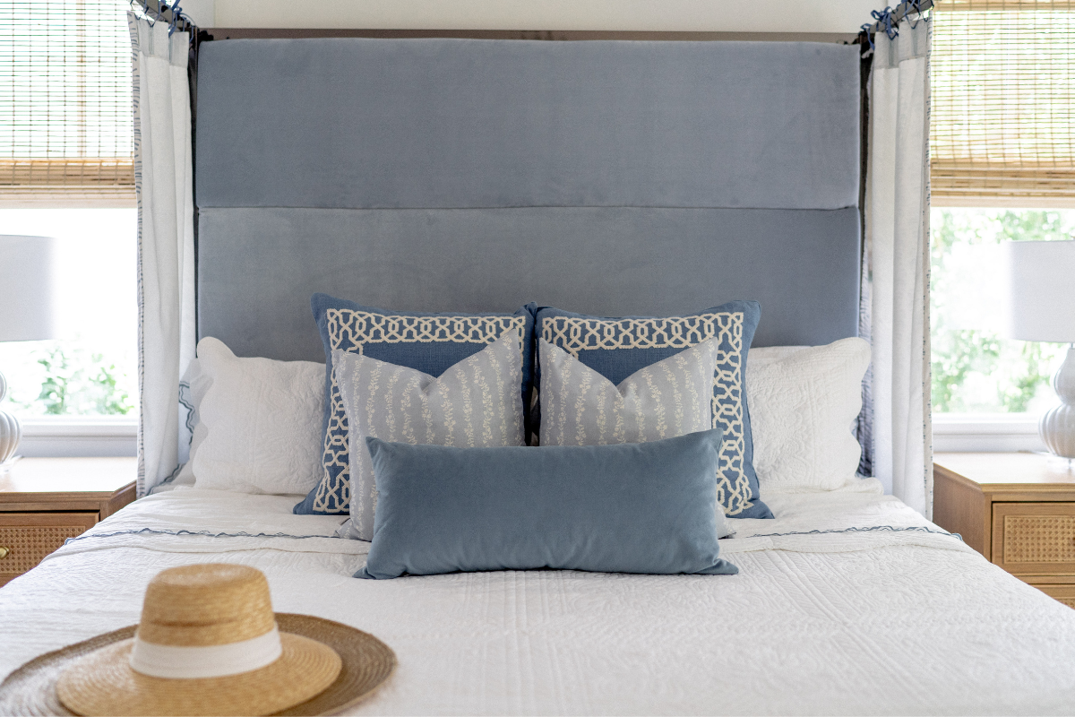 White Canopy Bed with a sky blue velvet headboard styled with Sky Blue Botanical Stripe Custom Curtains and four printed pillows by one blue velvet lumbar pillow on bed next to a summer hat laying on the edge