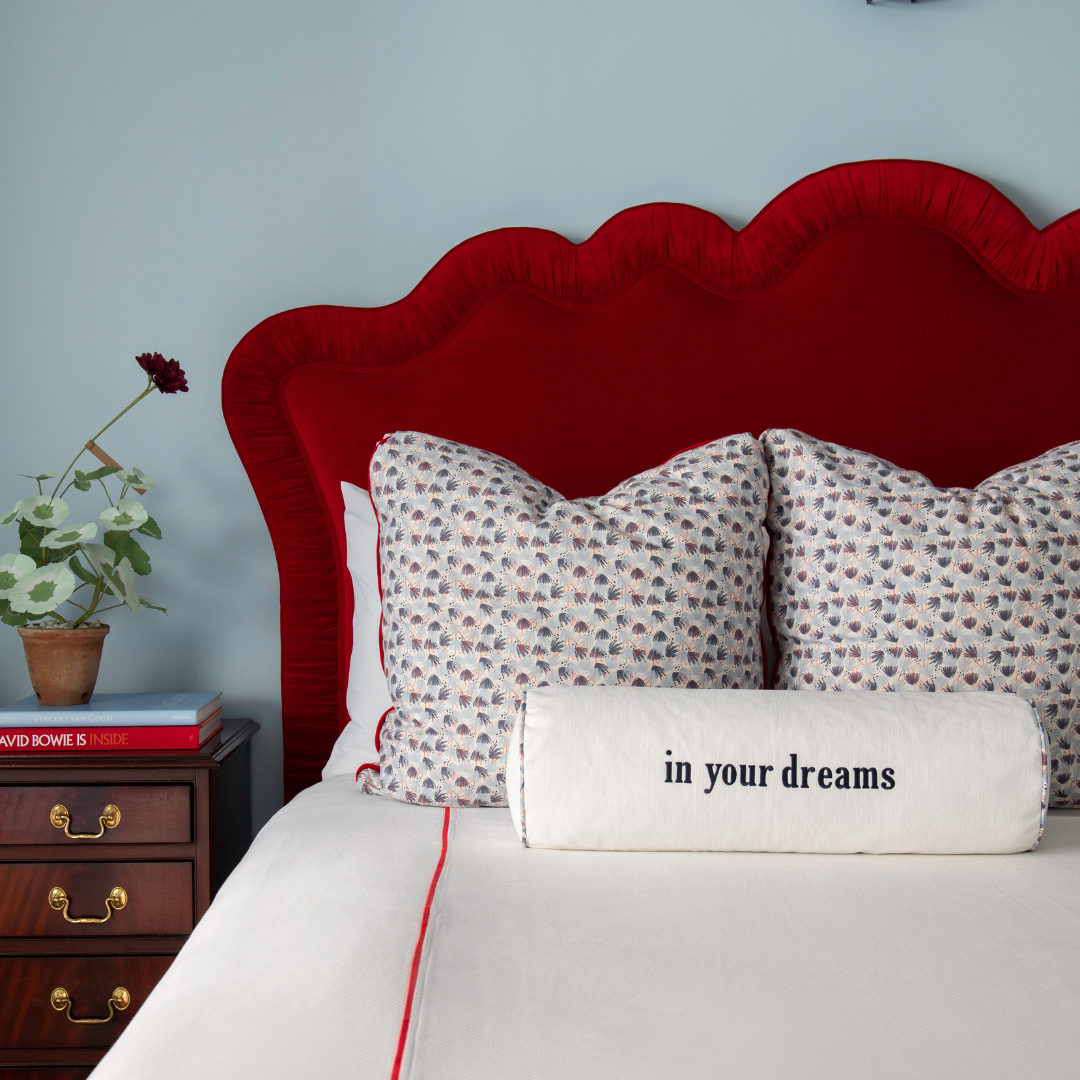 white bolster pillow on a bed embroidered with "in your dreams" paired with red and blue patterned pillows in a blue painted room