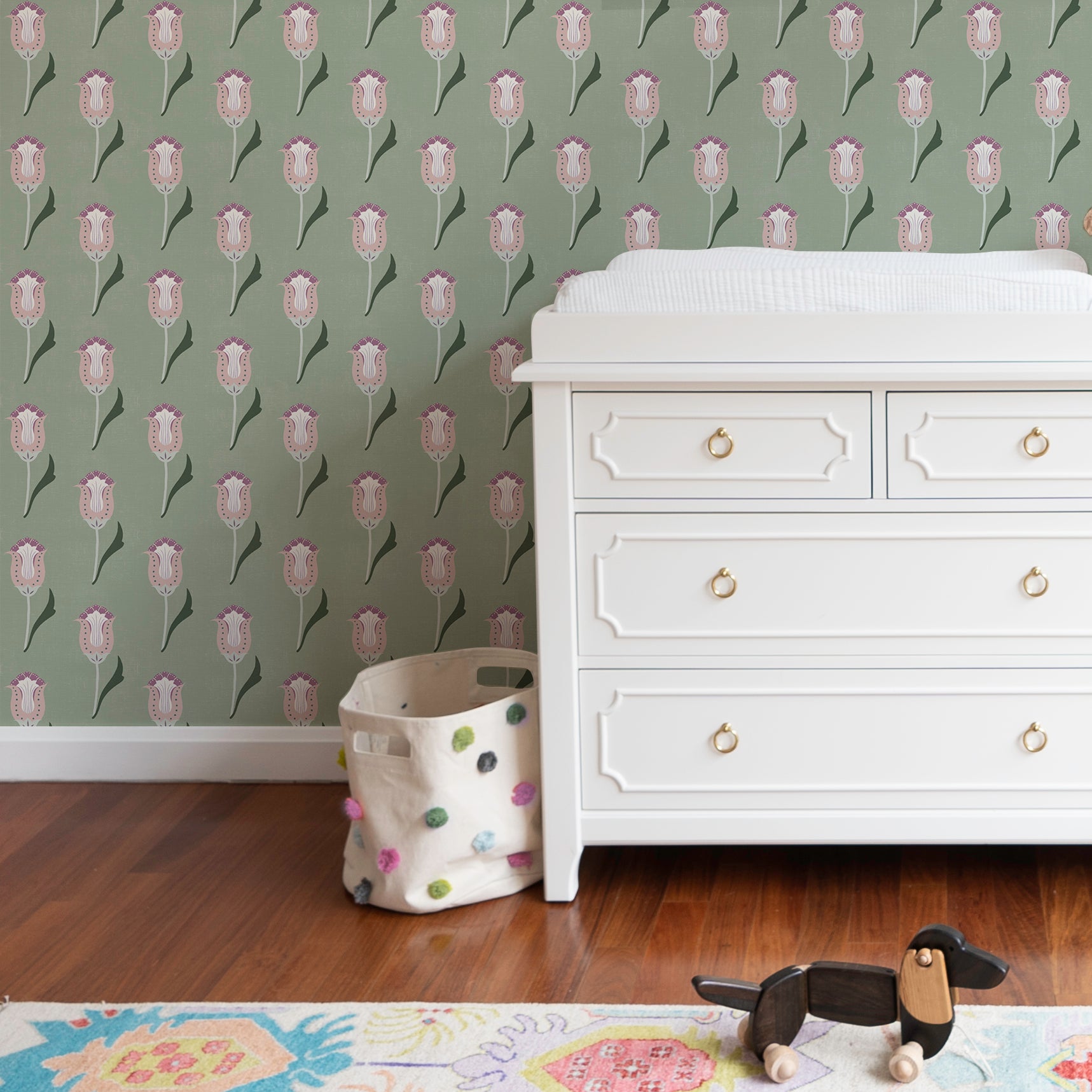 Close up of a nursery corner with a white changing table and green and pink floral drop repeat clay coated wallpaper styled with a toy dog