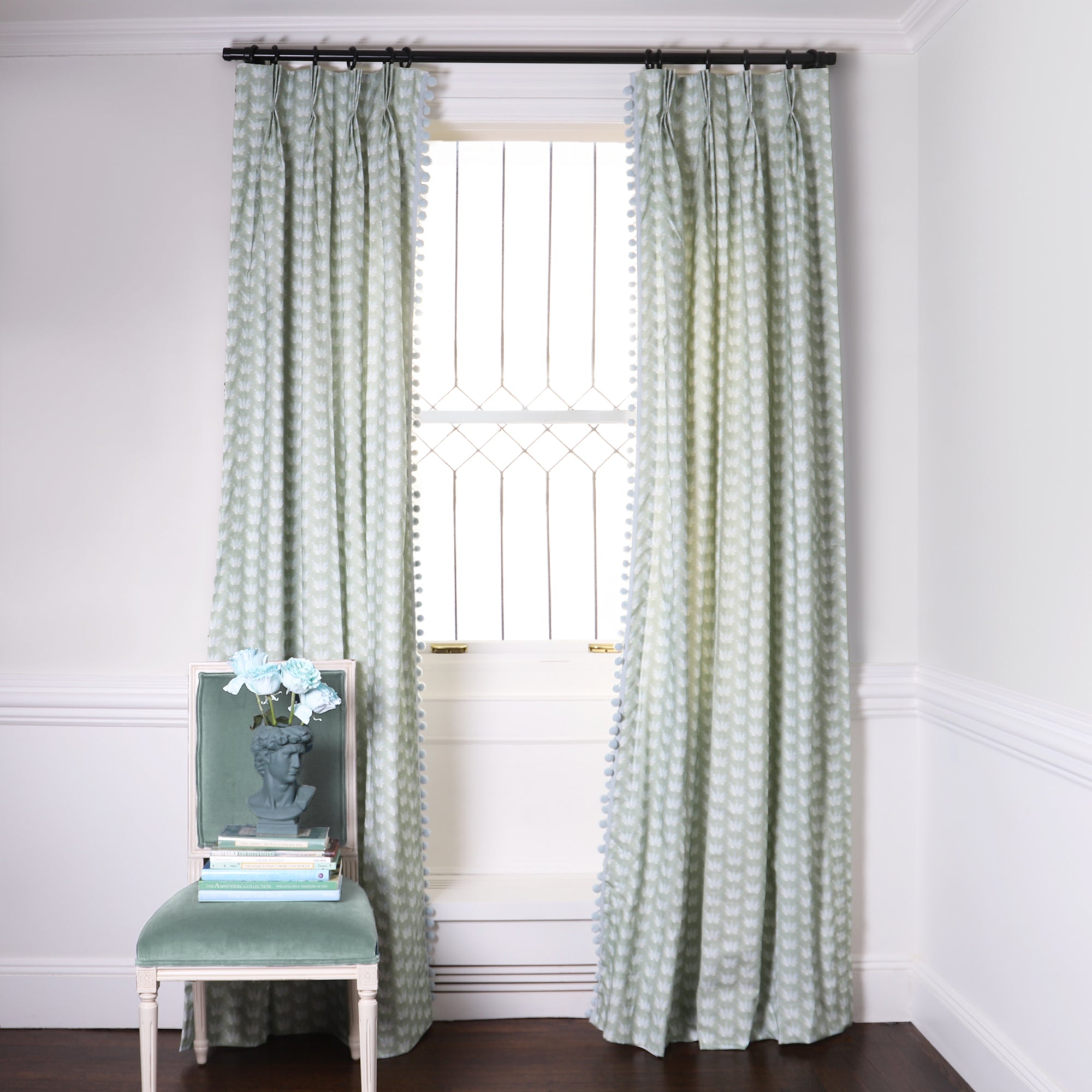 Blue & Green Floral Drop Repeat Custom Curtains on black rod in front of an illuminated window with a Blue Green Velvet chair with blue flowers in blue sculpted vase on top of stacked books