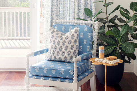 Close-up of white wooden chair with white and black printed pillow next to small table with a book and can on it next to a plant on a black claypot and an illuminated window styled with Sky Blue Botanical Stripe Custom Curtains