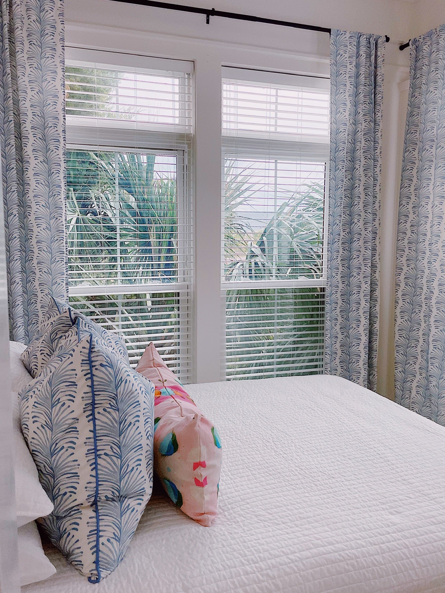 Sky blue botanical curtains hanging on a rod in front of a window in a bedroom with a white bed and sky blue botanical pillows