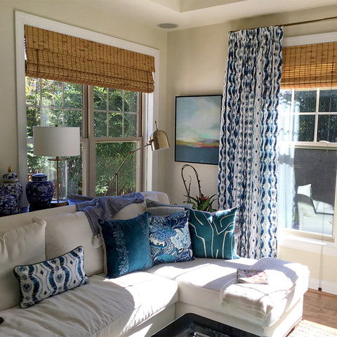 Living room corner with a white couch with three blue and white printed pillows on one side and a Blue Ikat Custom Pillow on the other side next to a window styled with Blue Ikat Stripe Custom Curtains