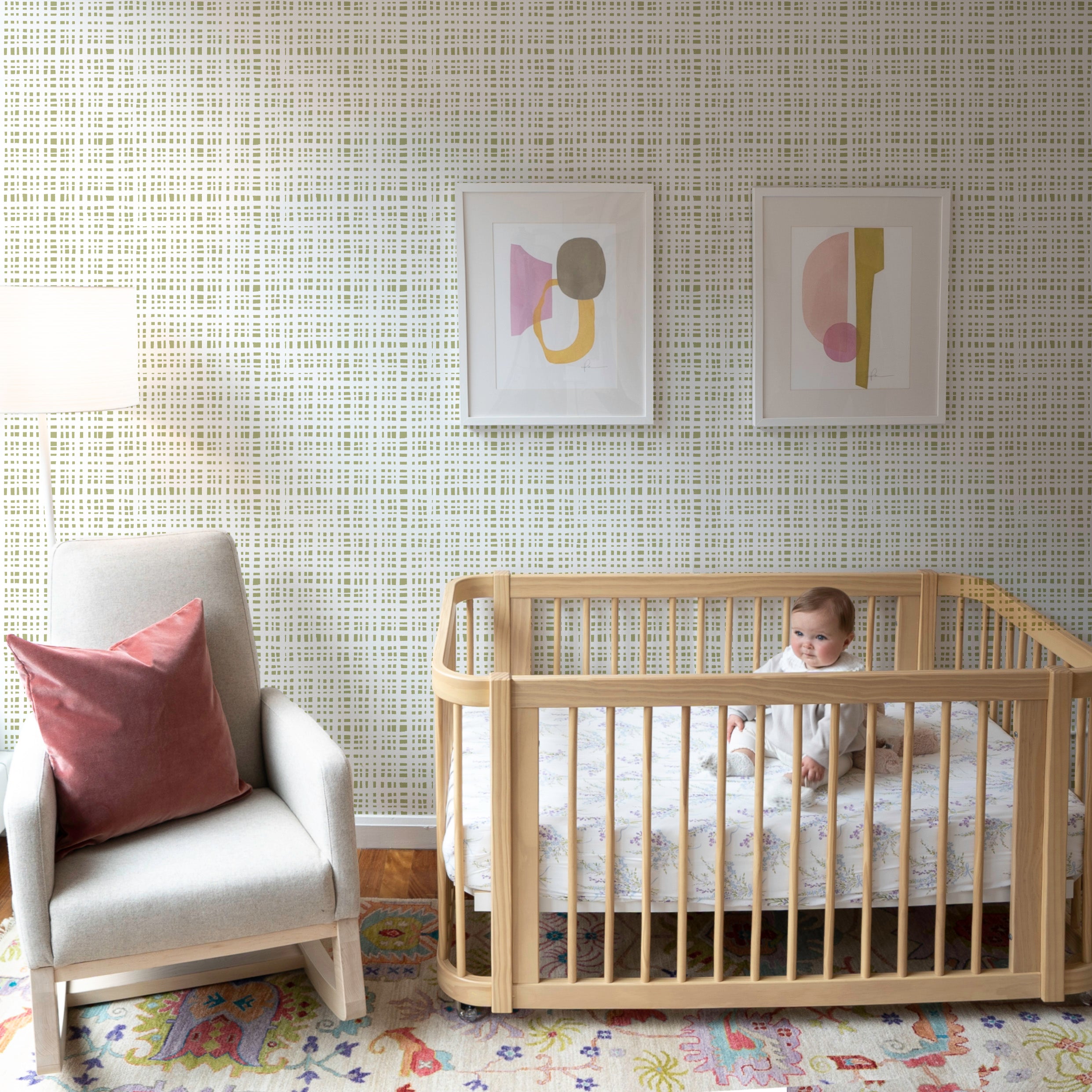 Nursery with plaid moss green clay coated wallpaper, a white glider and a wooden crib with a baby sitting up