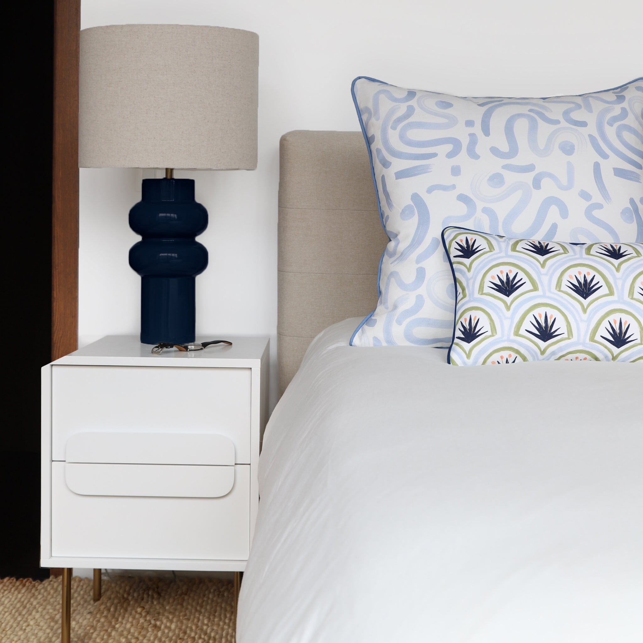 White Bed Close-up styled with a Sky Blue Custom Pillow and an Art Deco Palm Pattern Custom Lumbar Pillow next to white nightstand with navy and oat lamp on top