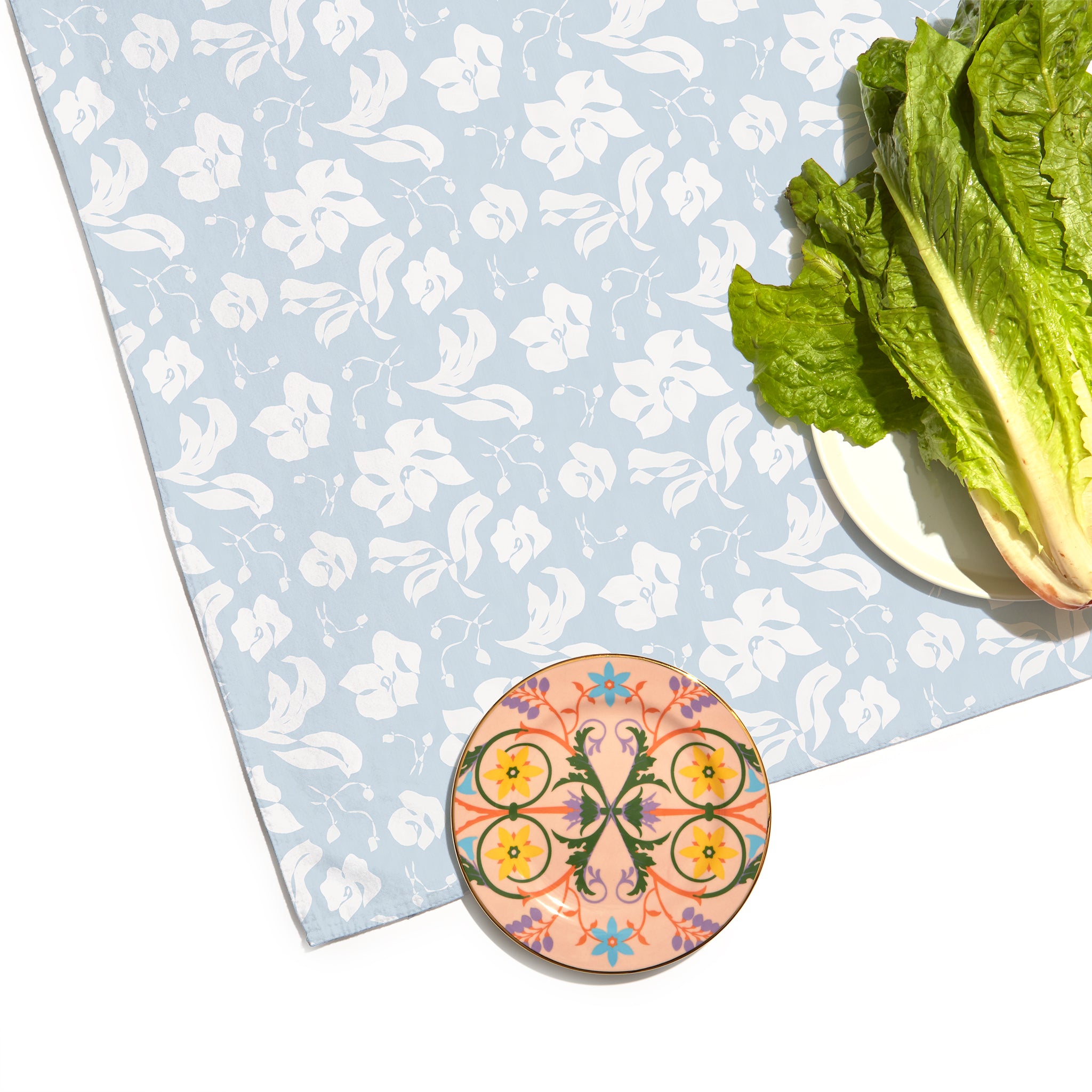 Corner Close-Up of Cornflower Blue Floral Custom Tablecloth with a colorful geometric shape printed plate and a white plate with lettuce on top