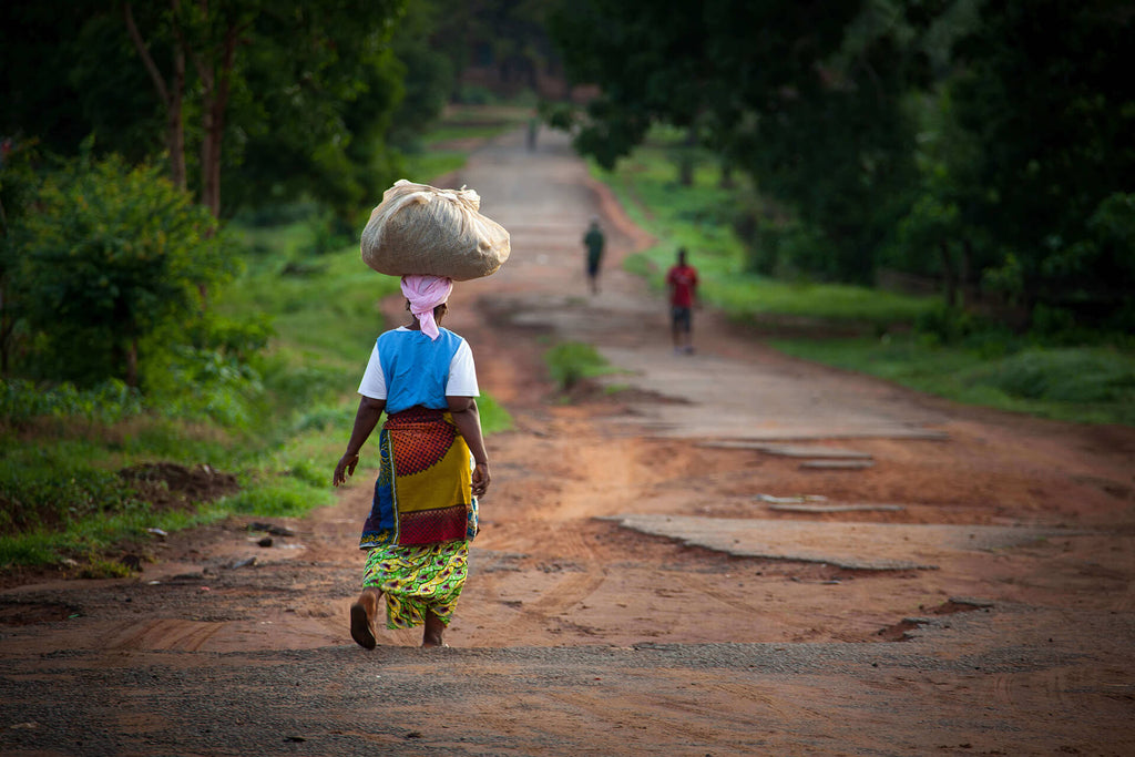 Woman from Sierra Leone in traditional clothing carrying a bag on top of her head and walking down the road.