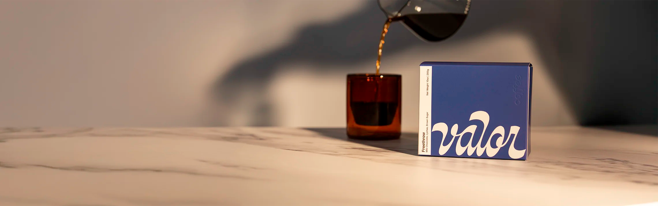 A box of Valor's signature blend, Freethrow, sits on a marble countertop. A glass carafe is seen pouring coffee into an amber colored glass mug.