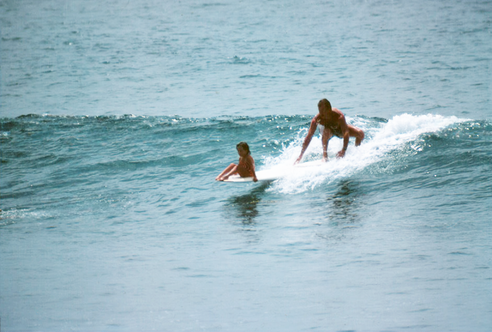 Bill Stewart surfing with daughter Ashley on the nose of his longboard, 1984