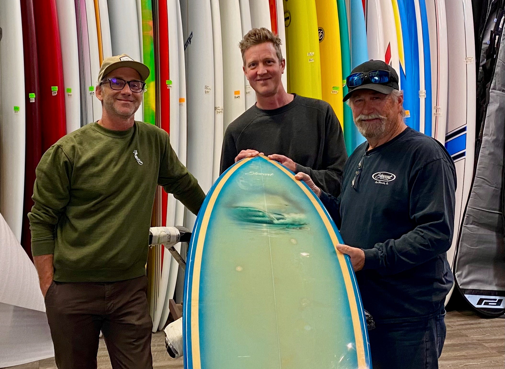 Benji Severson, Dan Severson, and Bill Stewart standing with an 80's twin fin surfboard