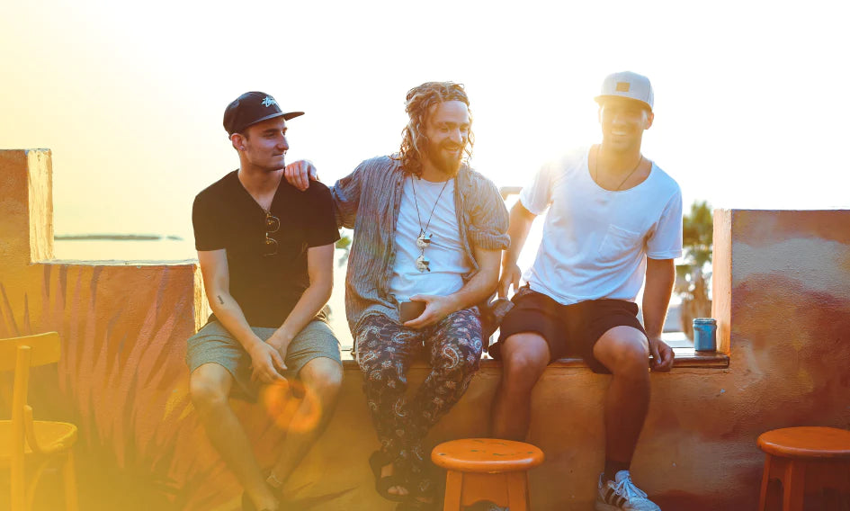 Three men sitting on stools outdoors, silhouetted against a colorful sunset sky.