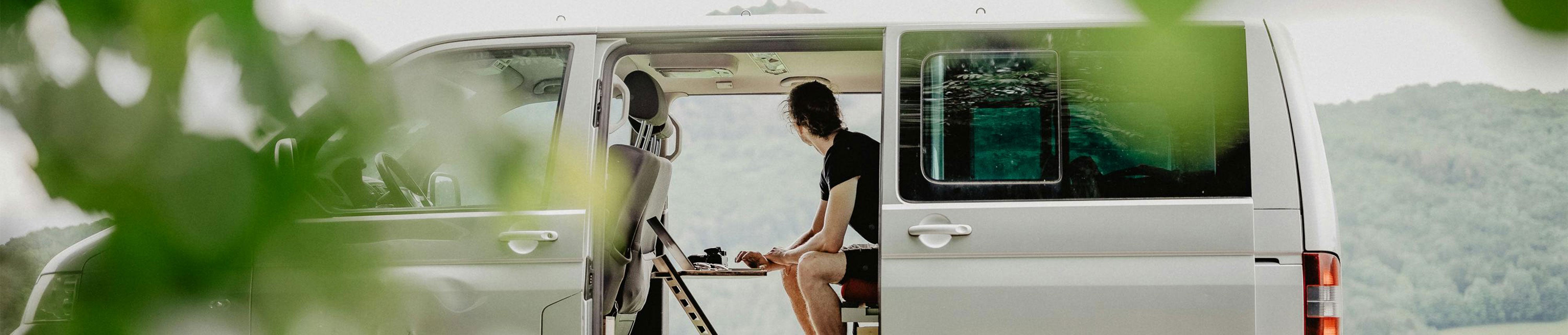 Person sitzt im hinteren Teil eines Vans an einem Standsome und blickt durch die offene Türe auf eine bergige Landschaft.