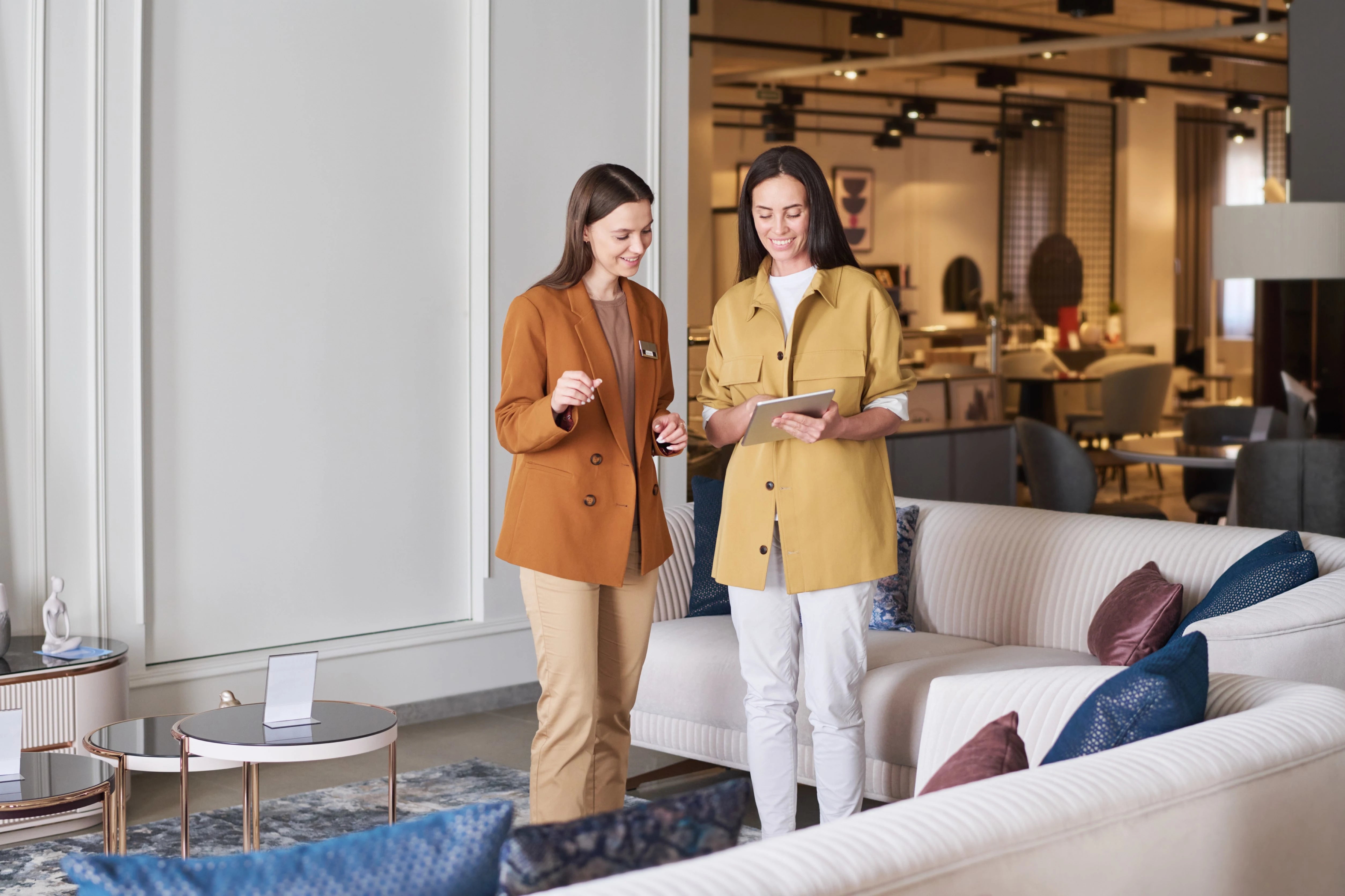Two female sales associates discussing design ideas on a tablet in a modern furniture showroom.