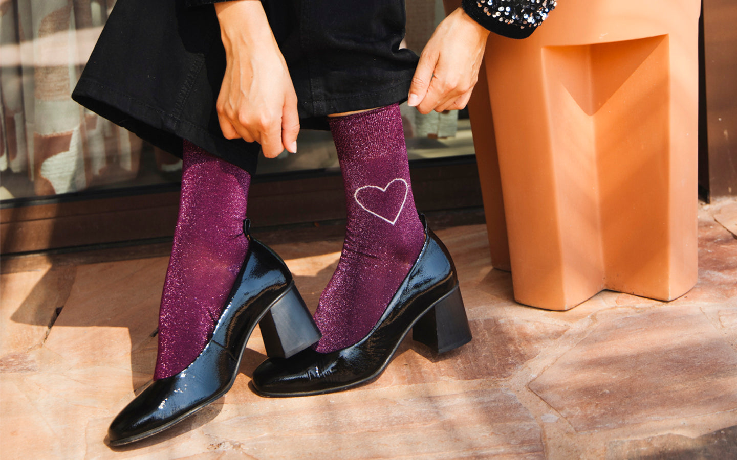 Zoom sur les pieds d’une mannequin portant des chaussettes en coton pailleté violet, ornées d’un cœur au contour blanc sur la cheville externe gauche. Elle porte des escarpins en vinyle noir et un pantalon noir qu’elle ajuste d’une main. La mannequin est assise sur un tabouret orangé, en extérieur sur une terrasse.