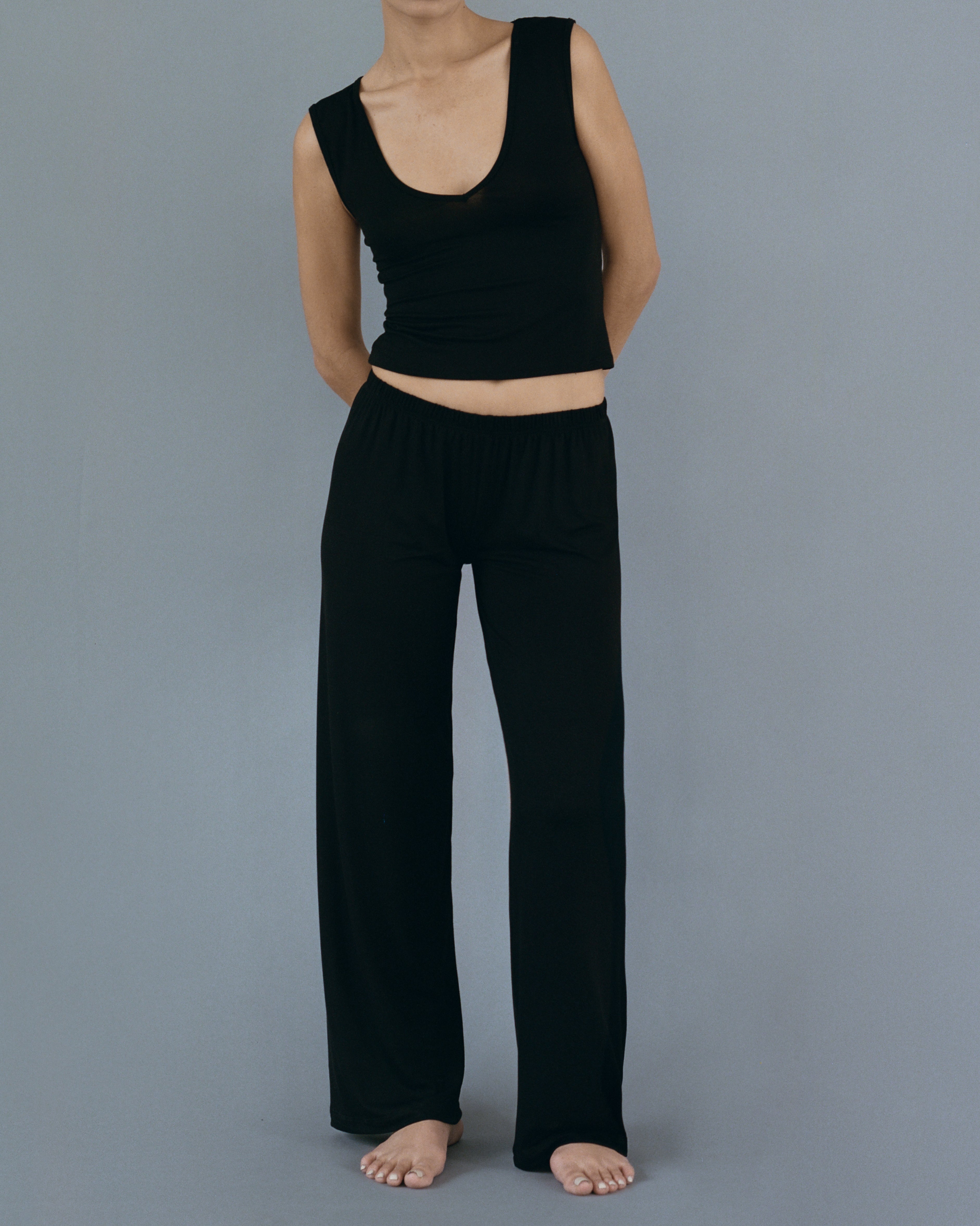 Model standing and wearing Kye Recline Shell bamboo v-neck fitted sleeveless tank in black. The tank’s fitted design and breathable bamboo fabric ensure comfort and style, showcasing the quality and craftsmanship of Kye Intimates.