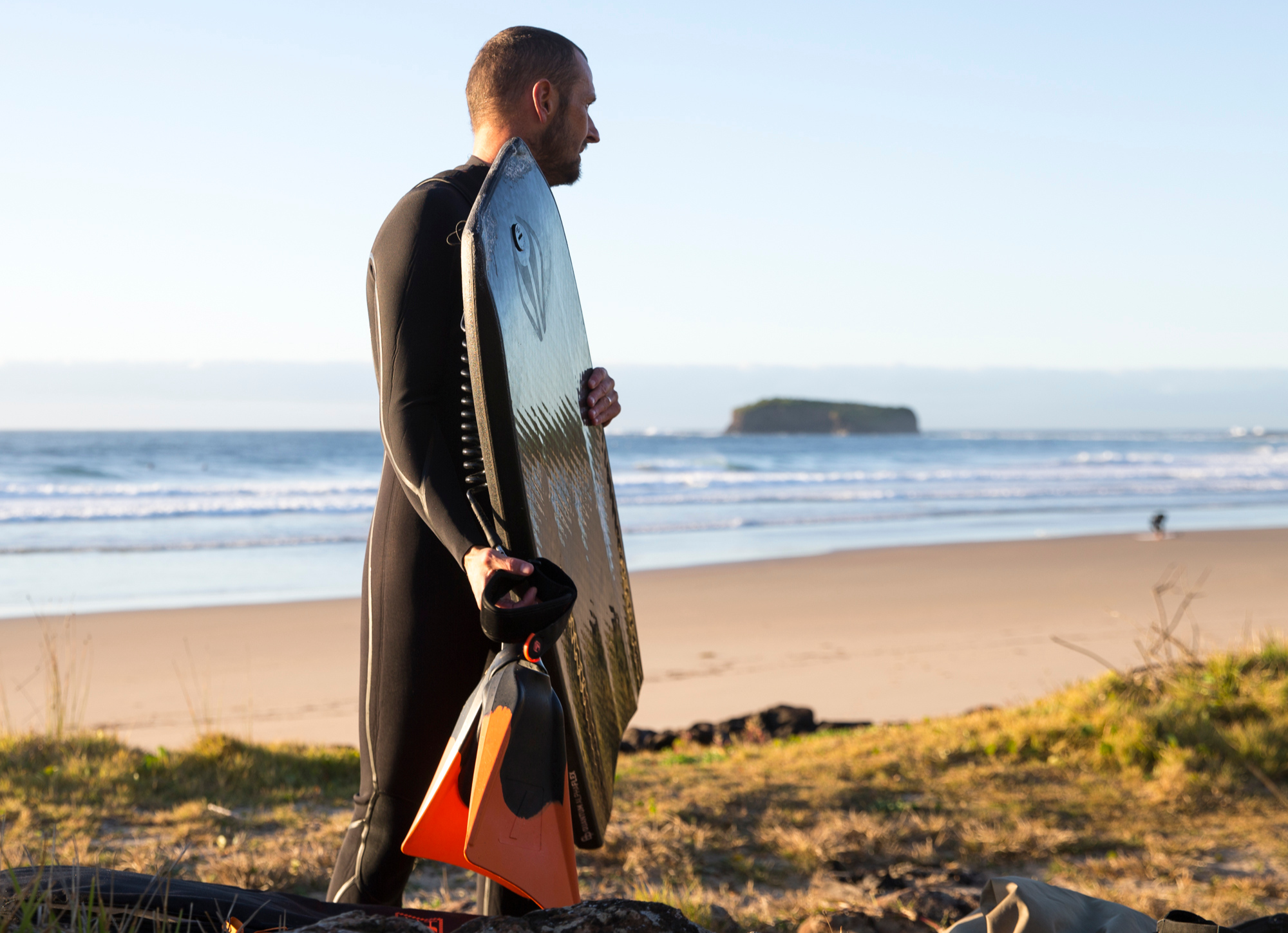 A bodyboarder holds swim fins and a board while looking out at waves.