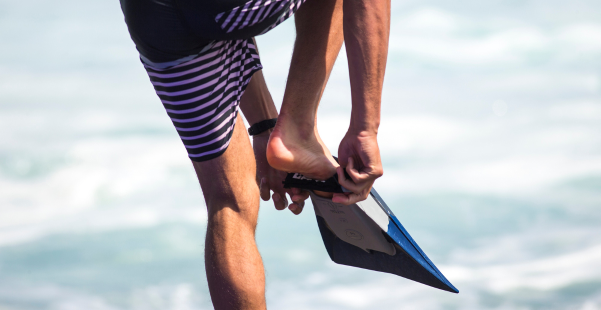 A bodyboarder puts a swim fin on his foot while standing on the beach.
