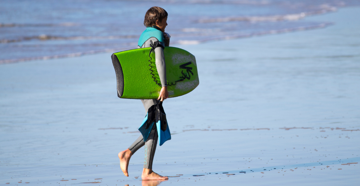 A kid walks along the shore while holding swim fins and a VS bodyboard.