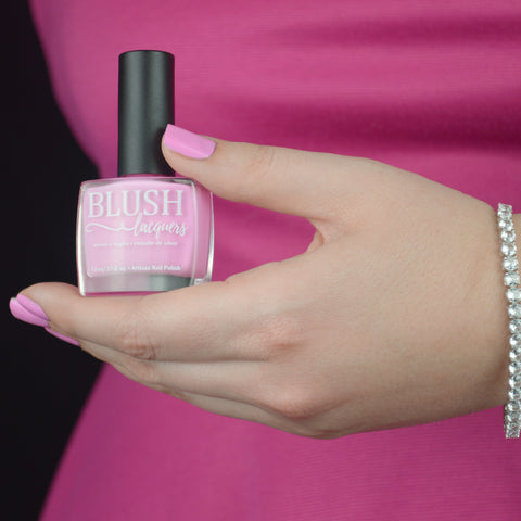 BLUSH Lacquers Malibu Beach Party cool toned baby pink creme nail polish being held by a woman's hand against a hot pink dress.