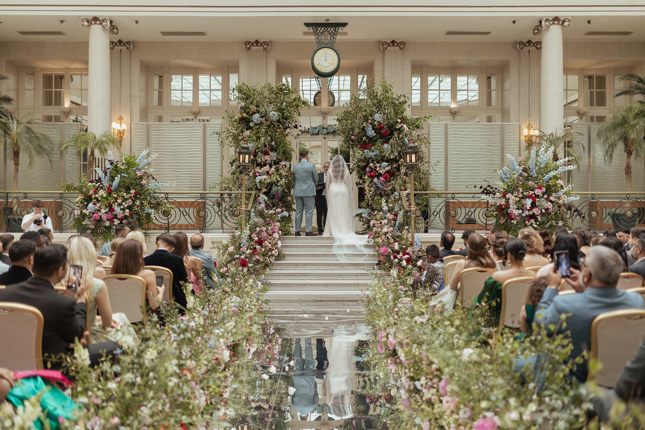  Bride and groom standing at the altar in The Waldorf Hilton's Palm Court in London, with opulent bespoke floral arrangements lining the grand staircase, reflecting the hotel’s blend of timeless elegance and modern allure - Amaranté London.