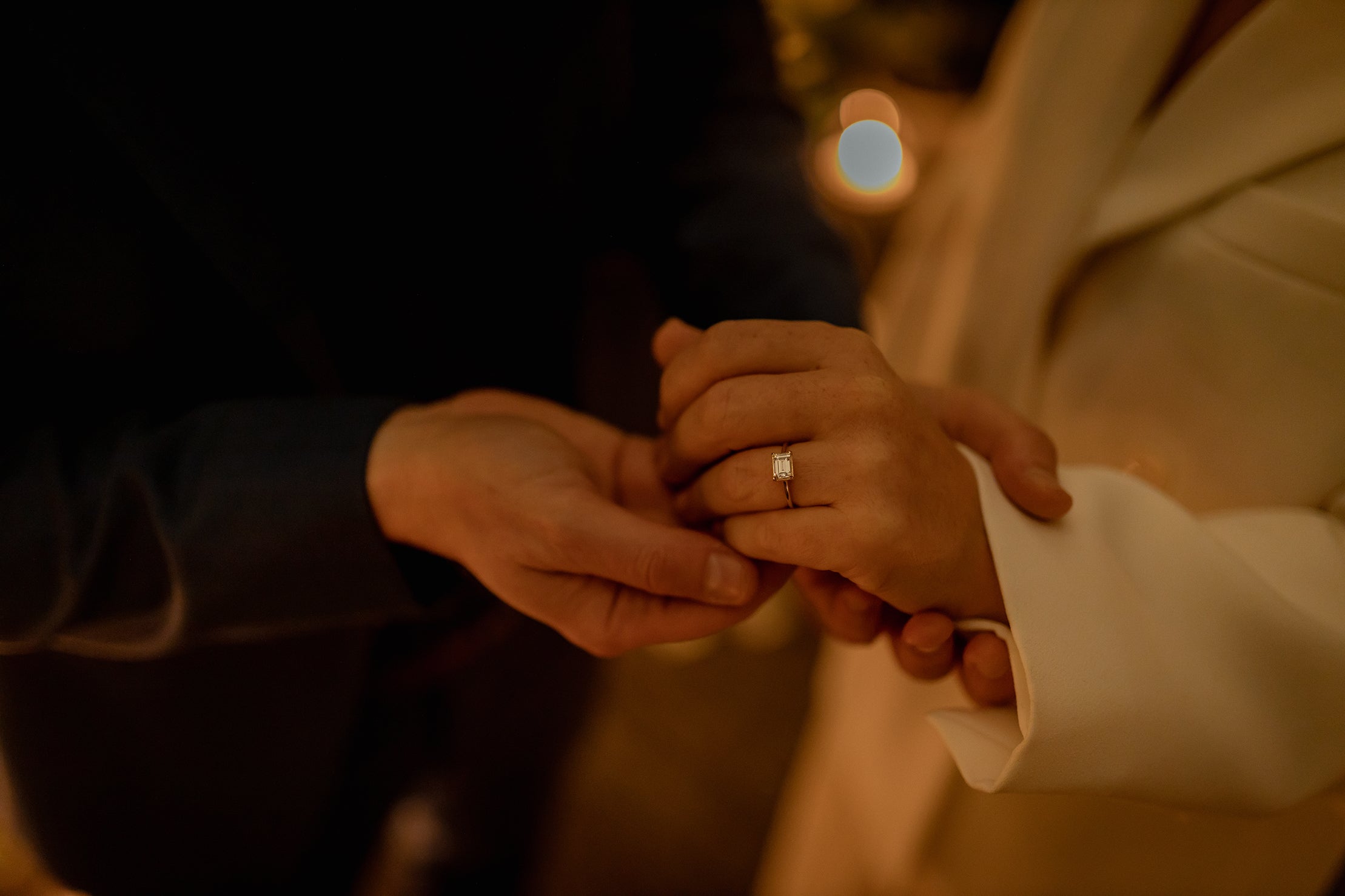 An intimate wedding proposal, a close-up of a couple's hands gently holding each other. The setting is intimate and dimly lit, with a warm golden glow illuminating the scene decorated with a bespoke floral installation designed and created by Amaranté London.