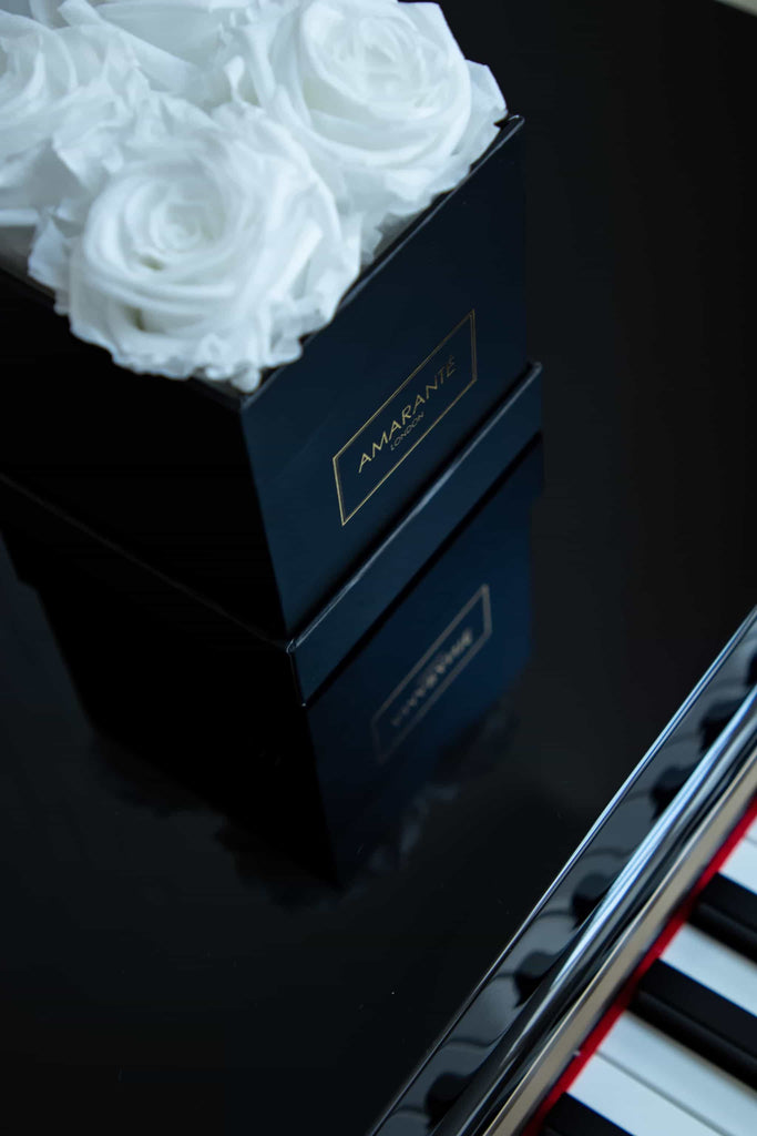 4 large white roses in a square black box with a matte finish, made with 100% recycled paper and cardboard, placed on the shiny black surface of a long tail piano