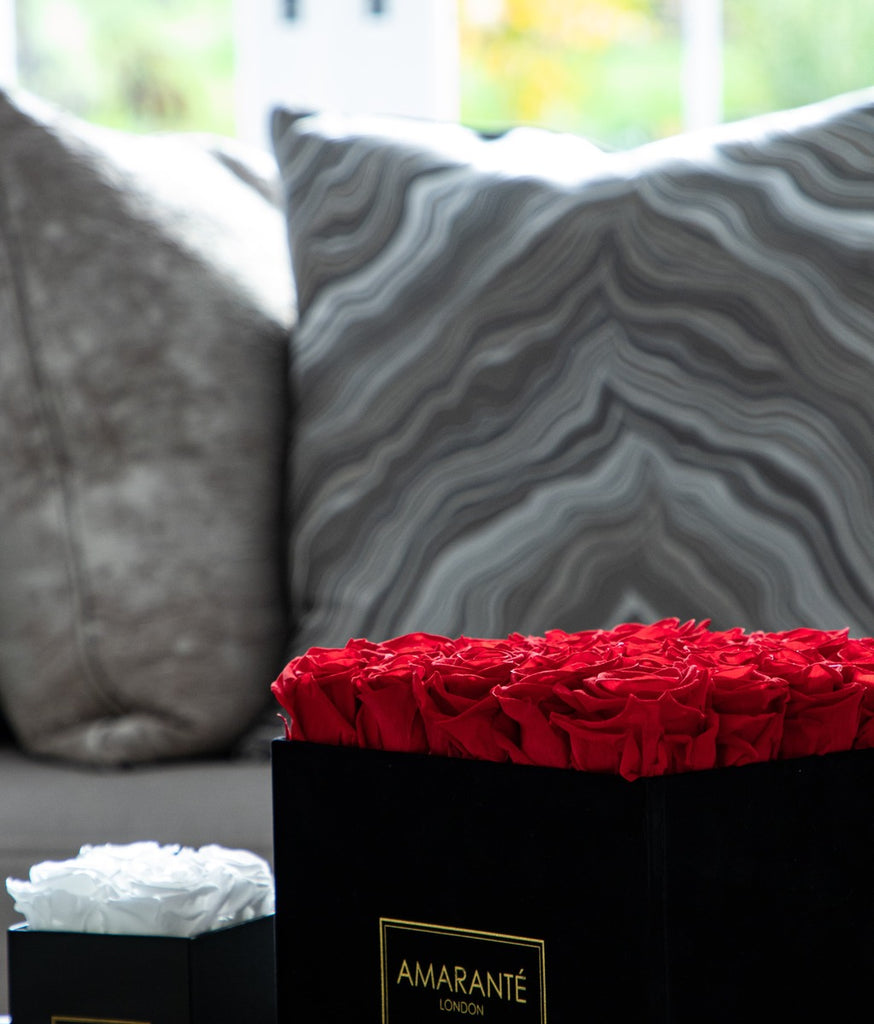A large square rose box with red roses along side a smaller one containing white roses on a coffe table in a living room.