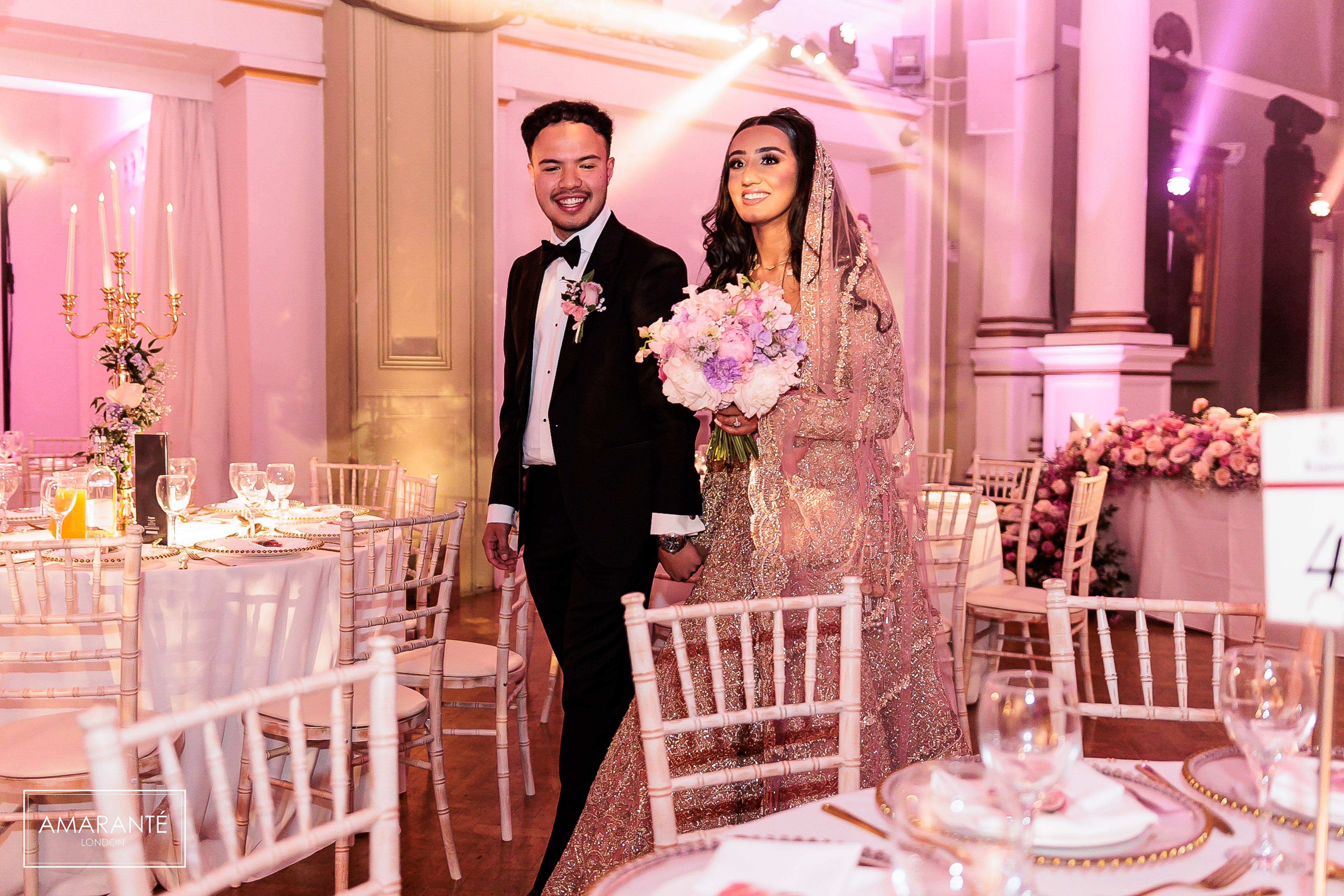 Joyous bride and groom are making their grand entrance into the reception hall, decorated with Amaranté bespoke floral installations. In a classic black tuxedo, the groom beams with pride next to the bride, who dazzles in a detailed, embellished gown with a bouquet of delicate wedding bouquet, showcasing Amaranté London's bespoke touch and experience in designing wedding installations.