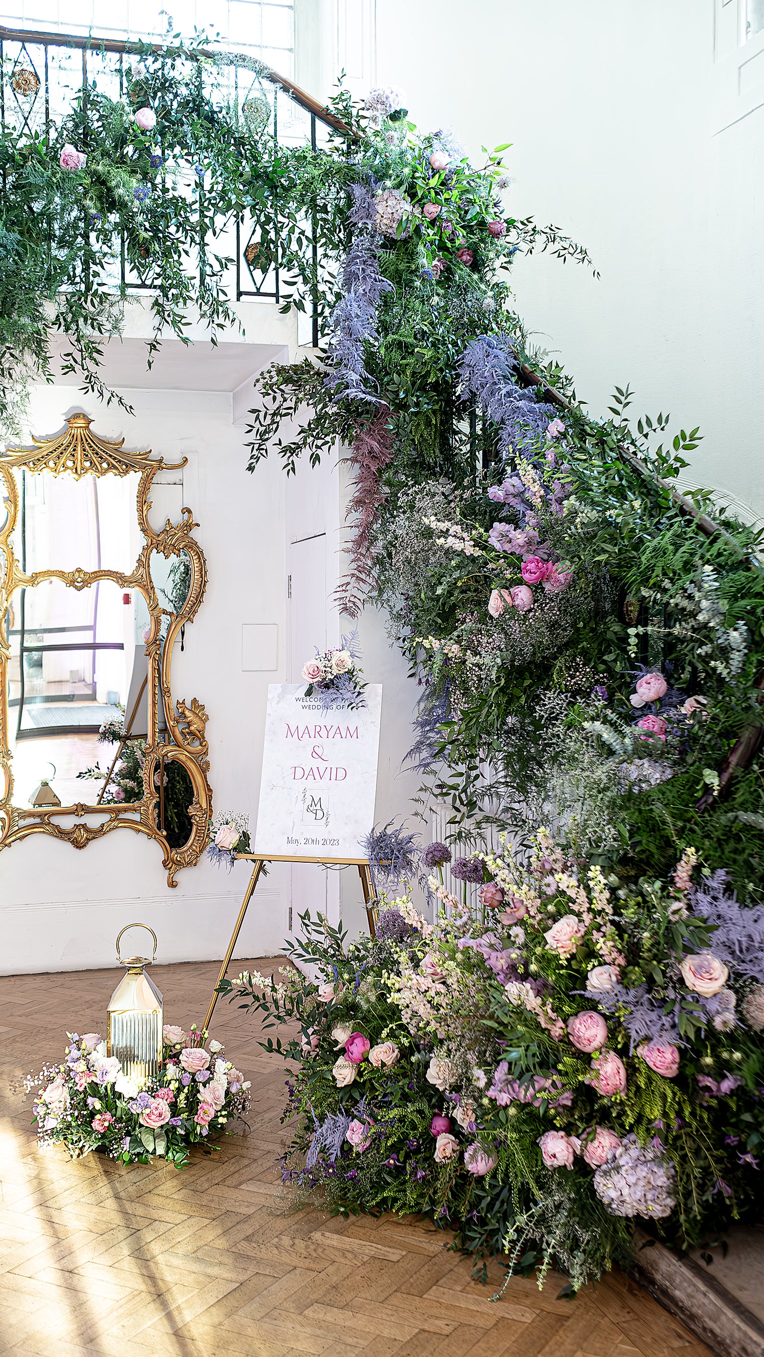 Elegant bespoke wedding floral installations frame the welcome sign at Maryam and David's wedding, designed by Event Florist Amaranté London. A lush arrangement of pastel roses, lilacs, and greenery cascades down the staircase, creating an enchanting entryway that promises a day filled with beauty and celebration.