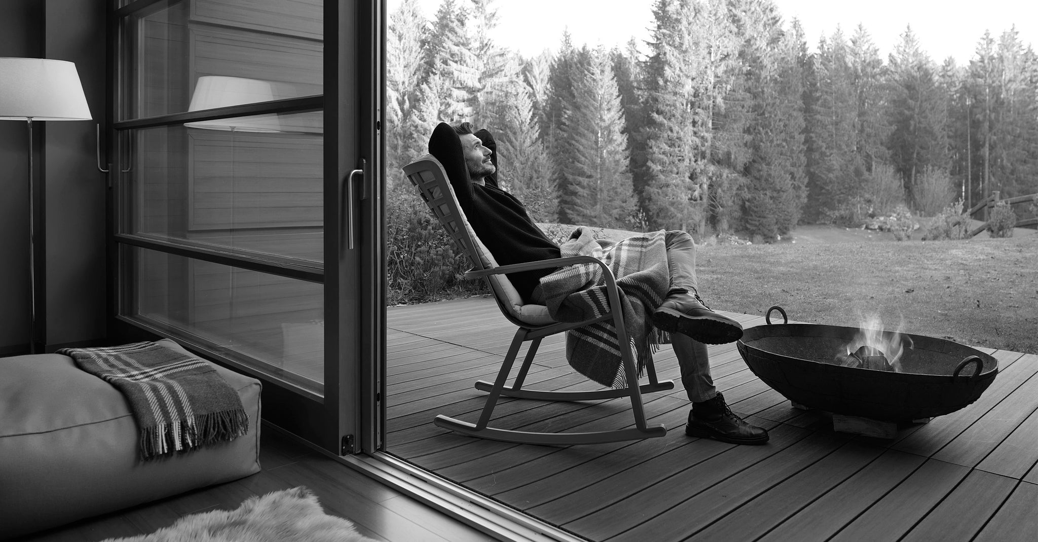 Un homme se détend dans un fauteuil à bascule Folio Rocking sur une terrasse avec cheminée et vue sur une clairière à la lisière de la forêt