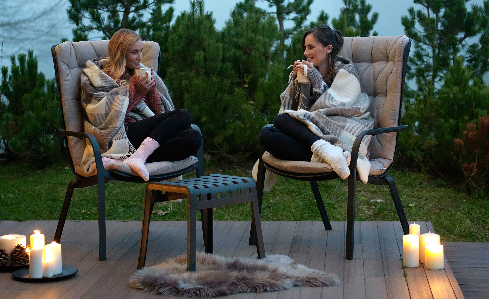 Deux filles sur des fauteuils Folio avec des repose-pieds Poggio enveloppés dans des couvertures sur la terrasse le soir