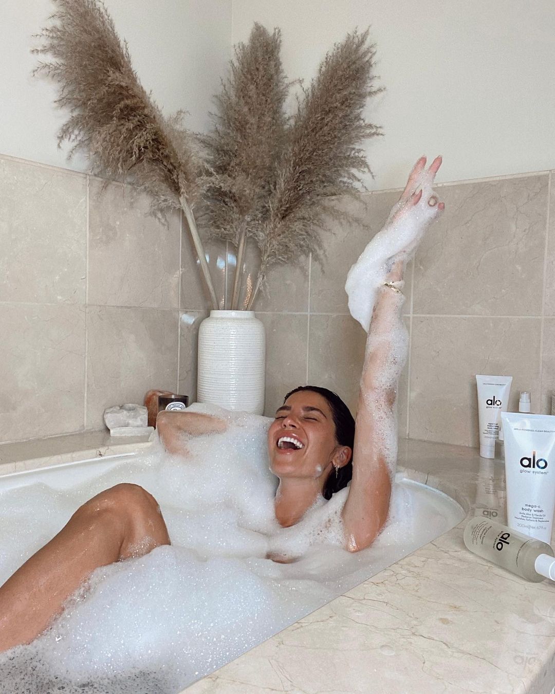 Femme aux cheveux foncés dans un bain moussant en marbre beige riant entourée de produits Alo Glow System.