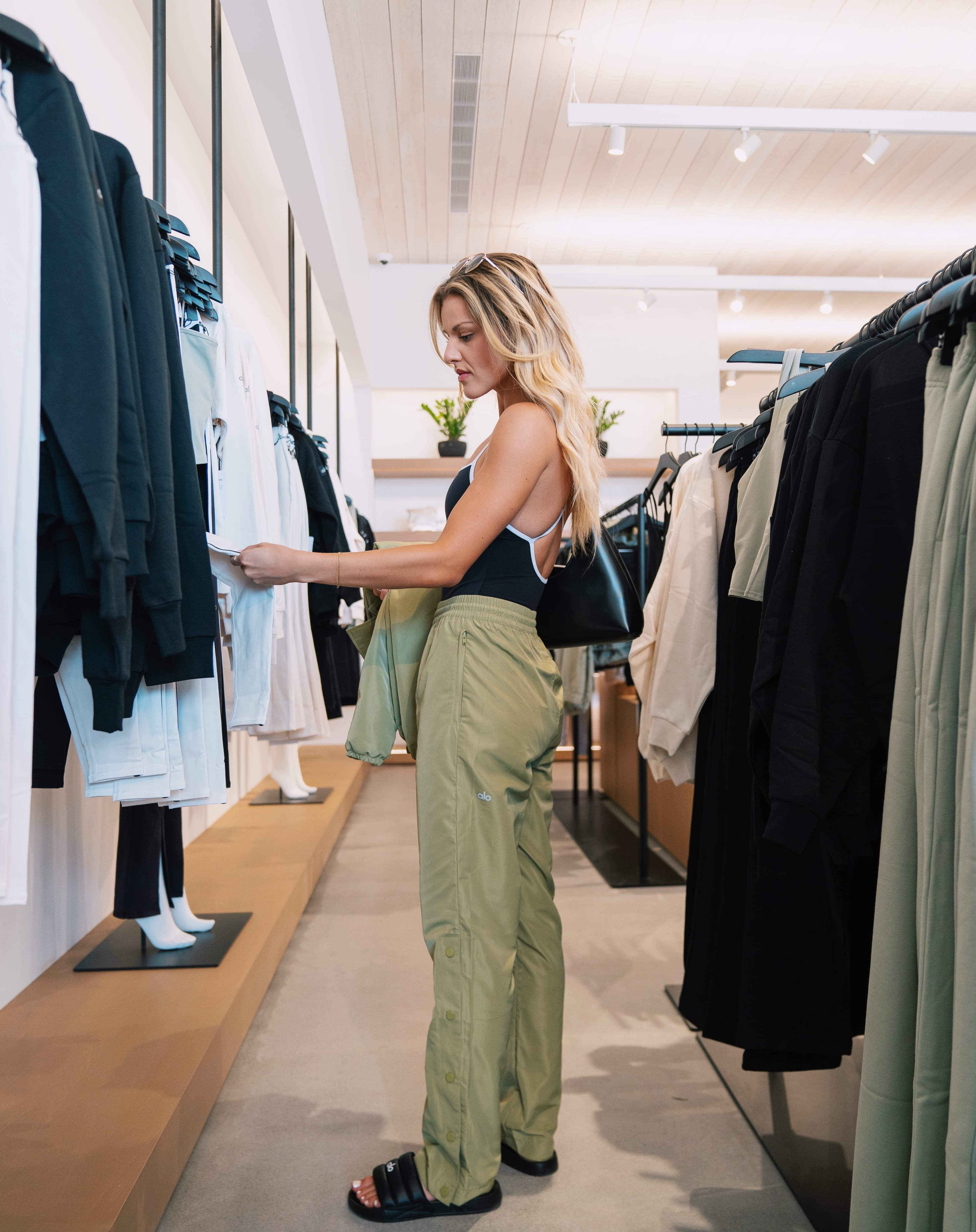 A woman shopping in an Alo Yoga Sanctuary wearing a pair of sage green pants with a snap side slits on the legs along with a pair of alo slides and bodysuit.