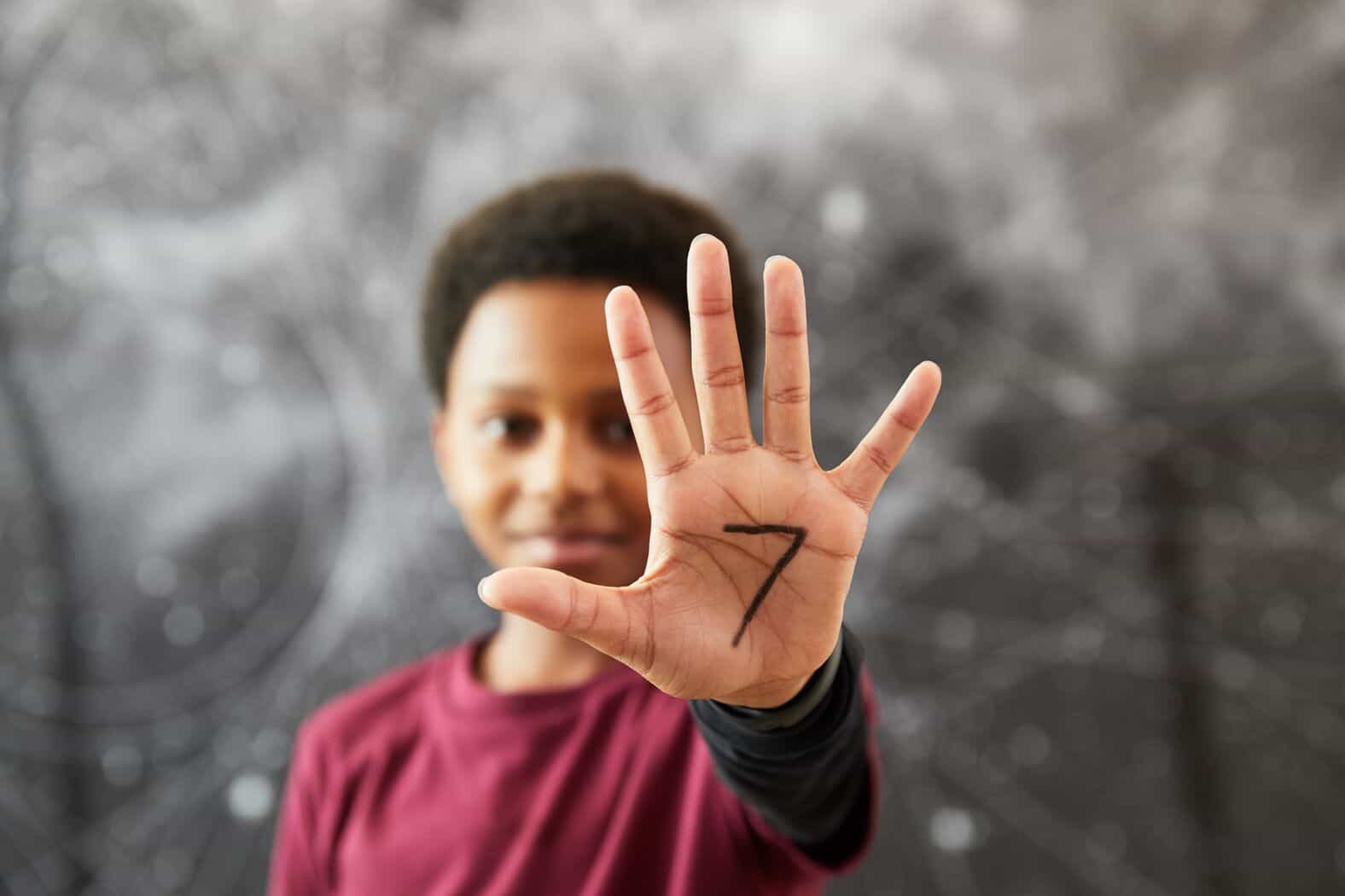 A young boy holding up the palm of his hand with the number 7 written on it to signify the age he was first introduced to yoga. 