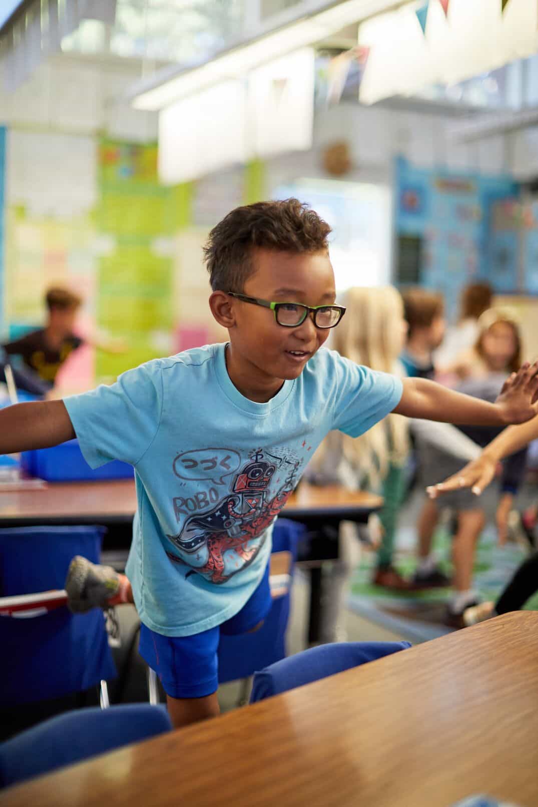 A young boy practicing yoga behind his desk in a classroom with his classmates.  