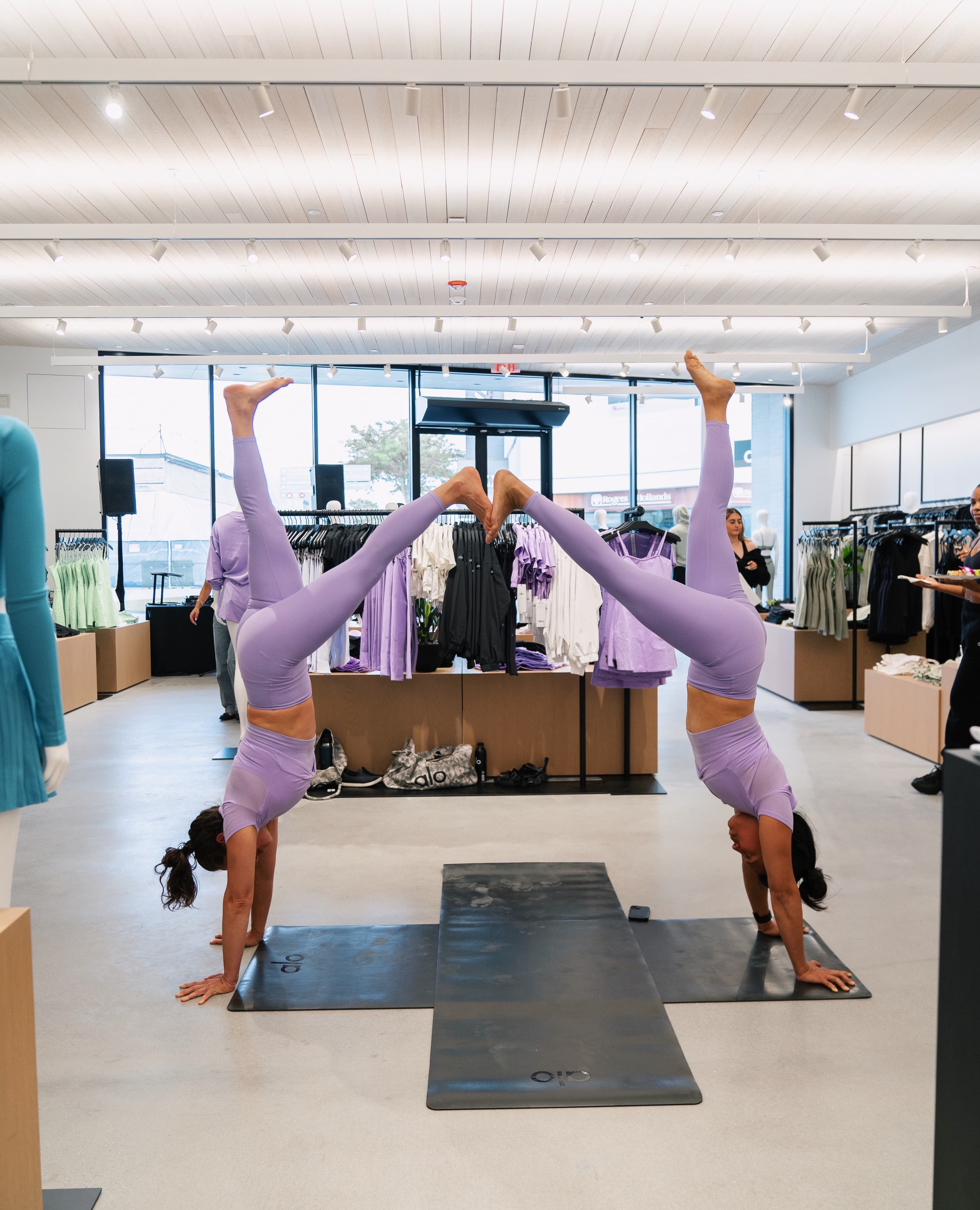 A photo of two women in handstands with one of each of their feet touching the other person’s in the middle of the Alo Old Orchard store wearing purple workout sets.  