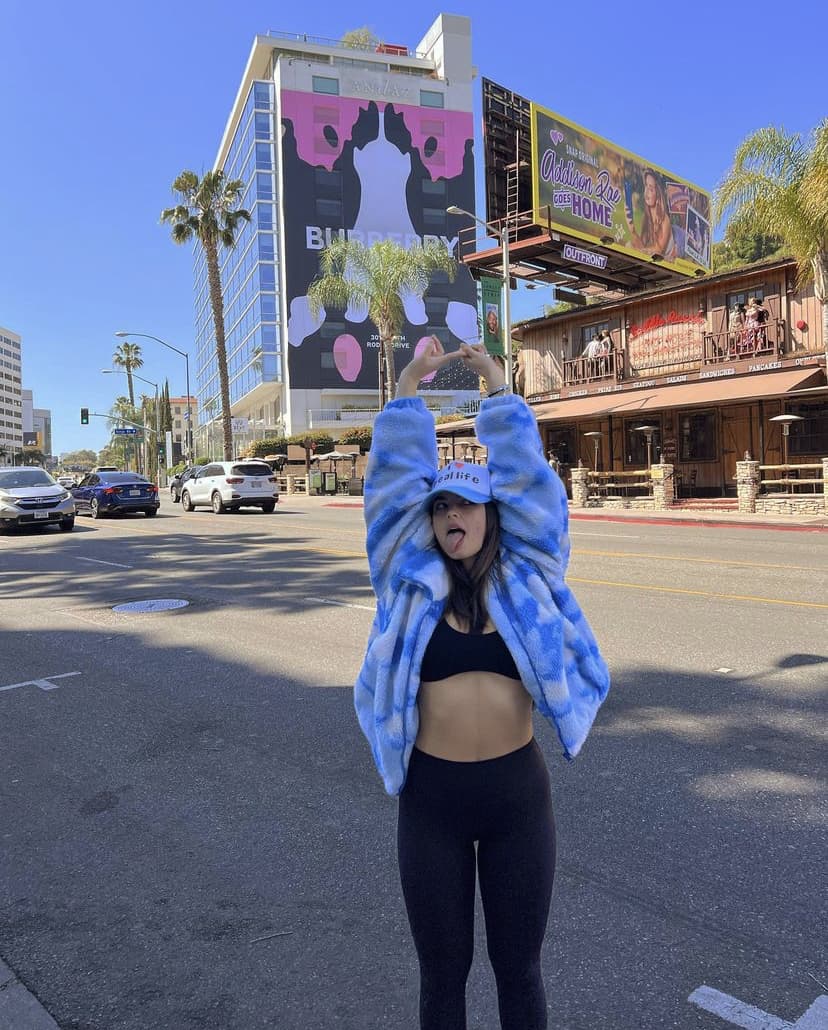 A photo of Addison Rae posing with her arms above her head and sticking her tongue out wearing a micro waffle sports bra and black high waist legged on a street on Southern California.  