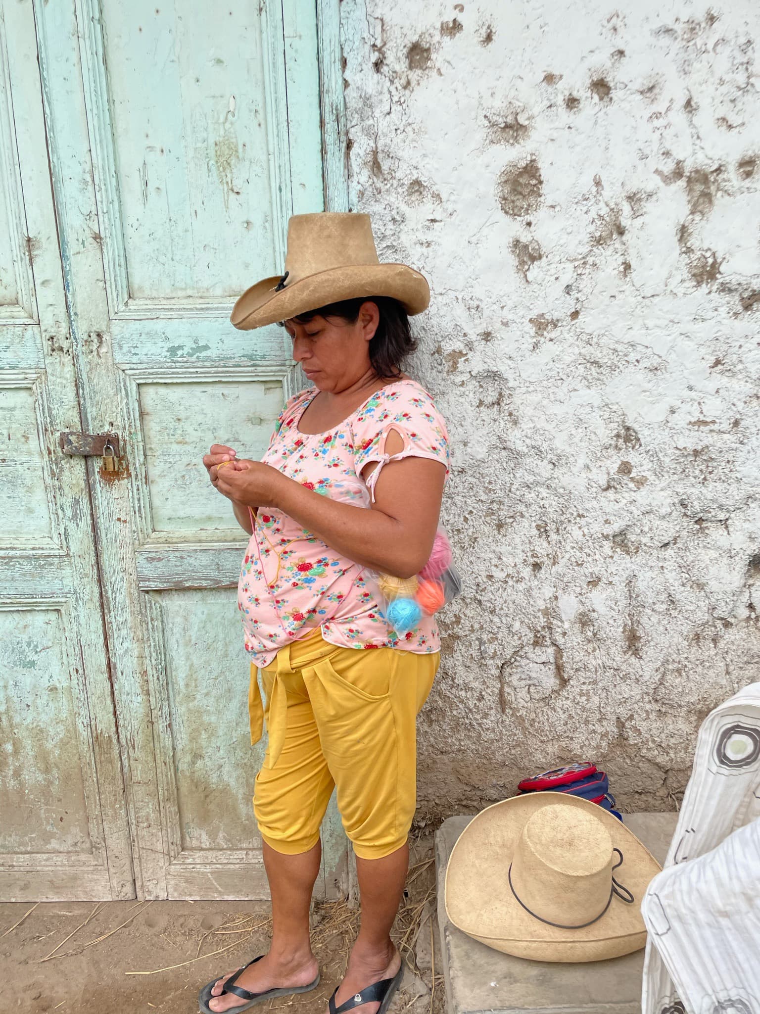 A woman wearing a traditional hat while weaving a crochet top standing against a pale teal door and concrete wall.  