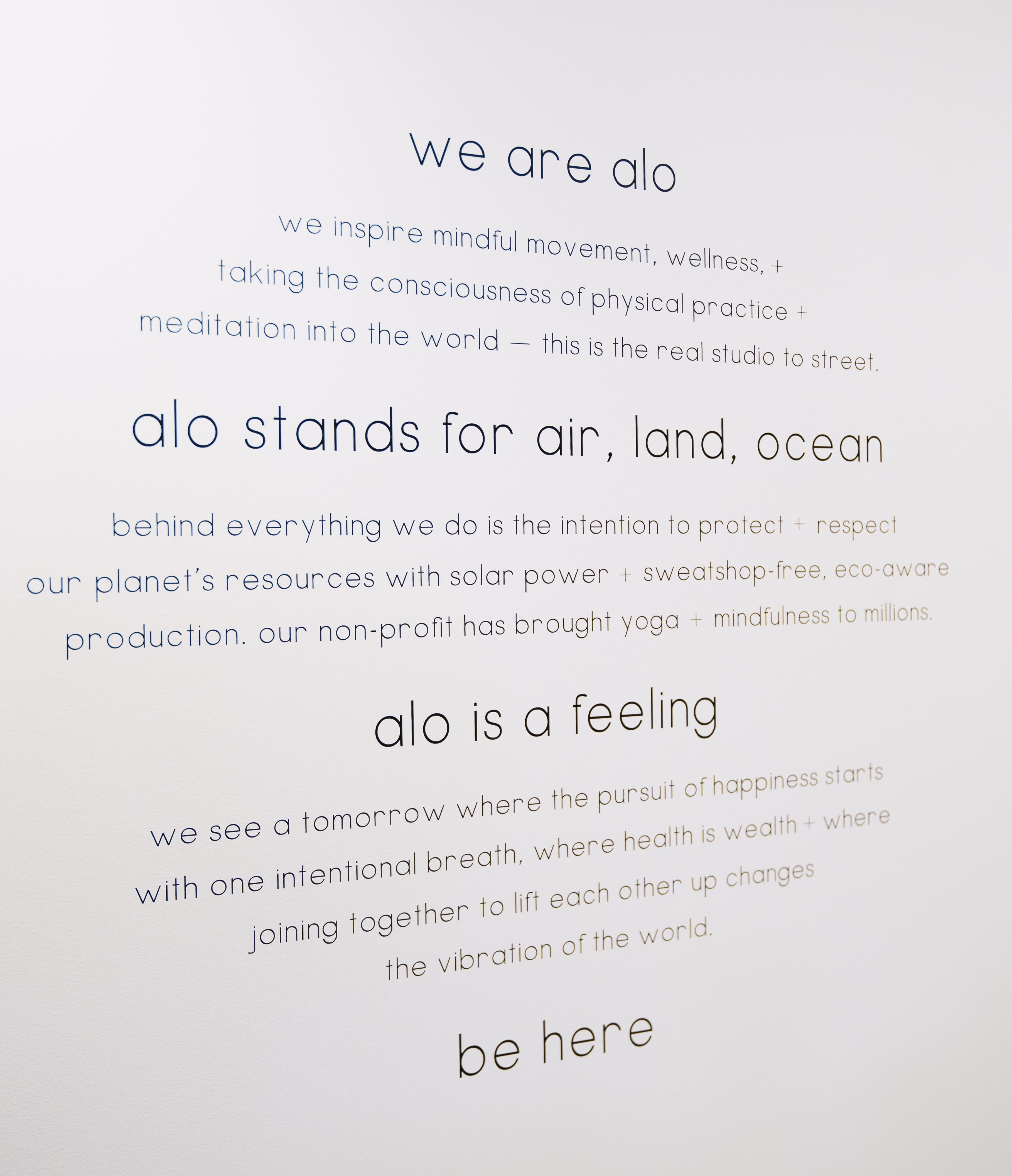 A photo of a wall sticker at the Alo Old Orchard store in Skokie Illinois with the headers, “We are Alo,” “Alo stands for air, land, ocean,” “Alo is a feeling,” and “be here” with sub-headers underneath that are slightly blurred.  