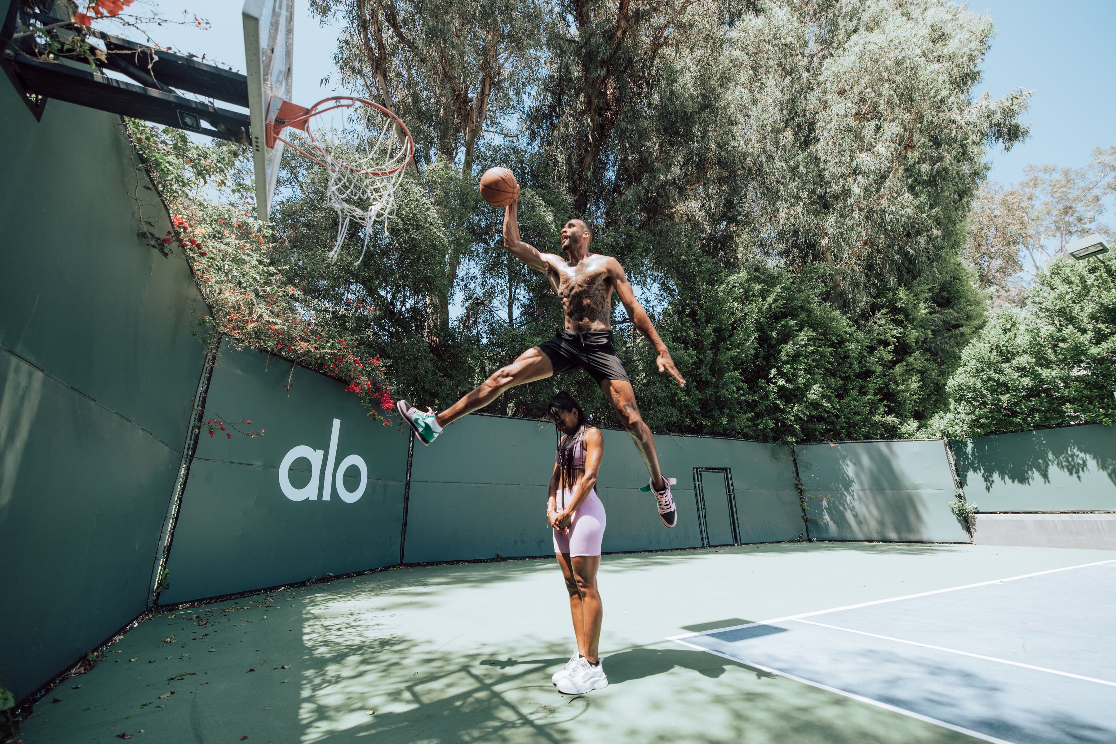 A photo of a man jumping over a woman to dunk a basketball on the court at Alo Summer House 2022.  