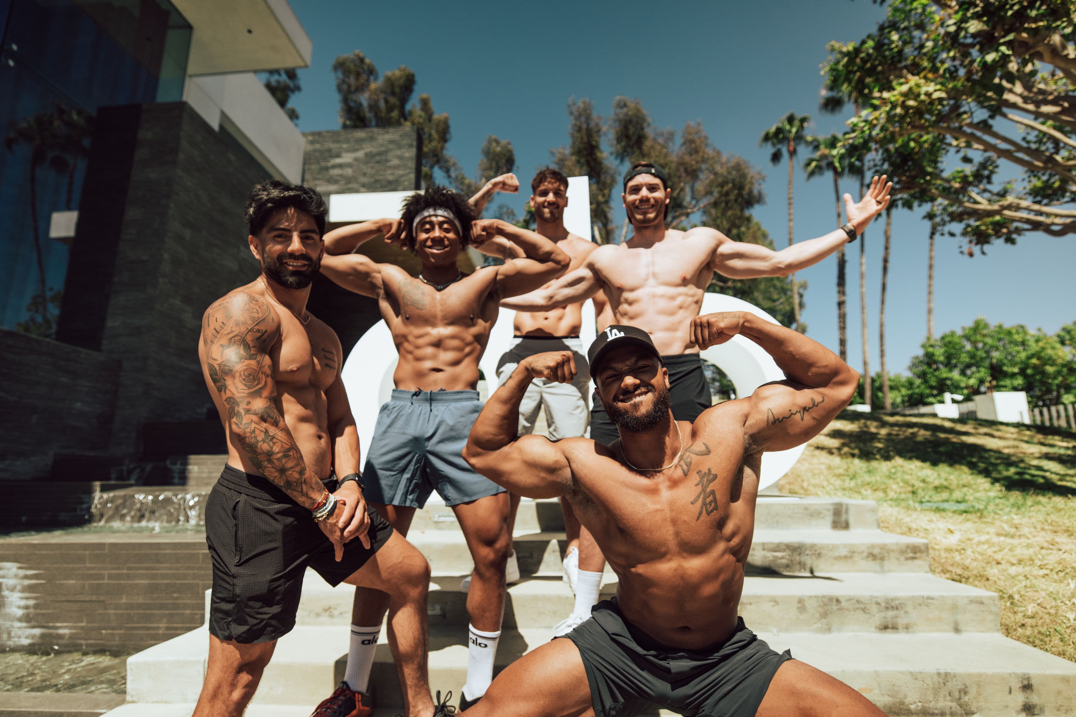 Five men posing on the stairs at Alo Summer House 2022 while all flexing at the camera wearing Alo Yoga shorts and no shirts. 