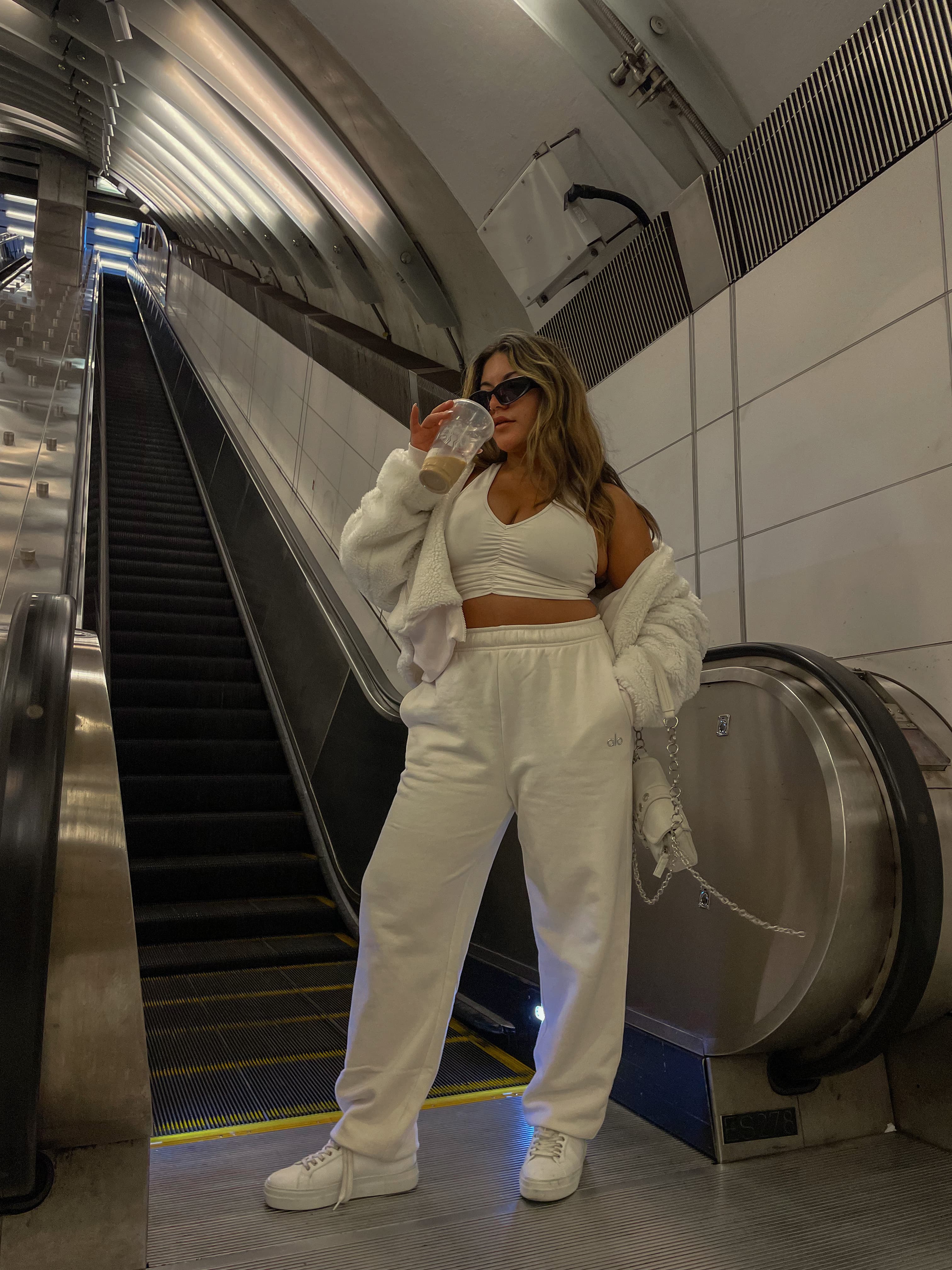 A woman wearing an all-white streetwear set with baggy sweatpants, a ruched sports bra, and a sherpa jacket draped over her shoulder while sipping a coffee standing at the bottom of an escalator.  